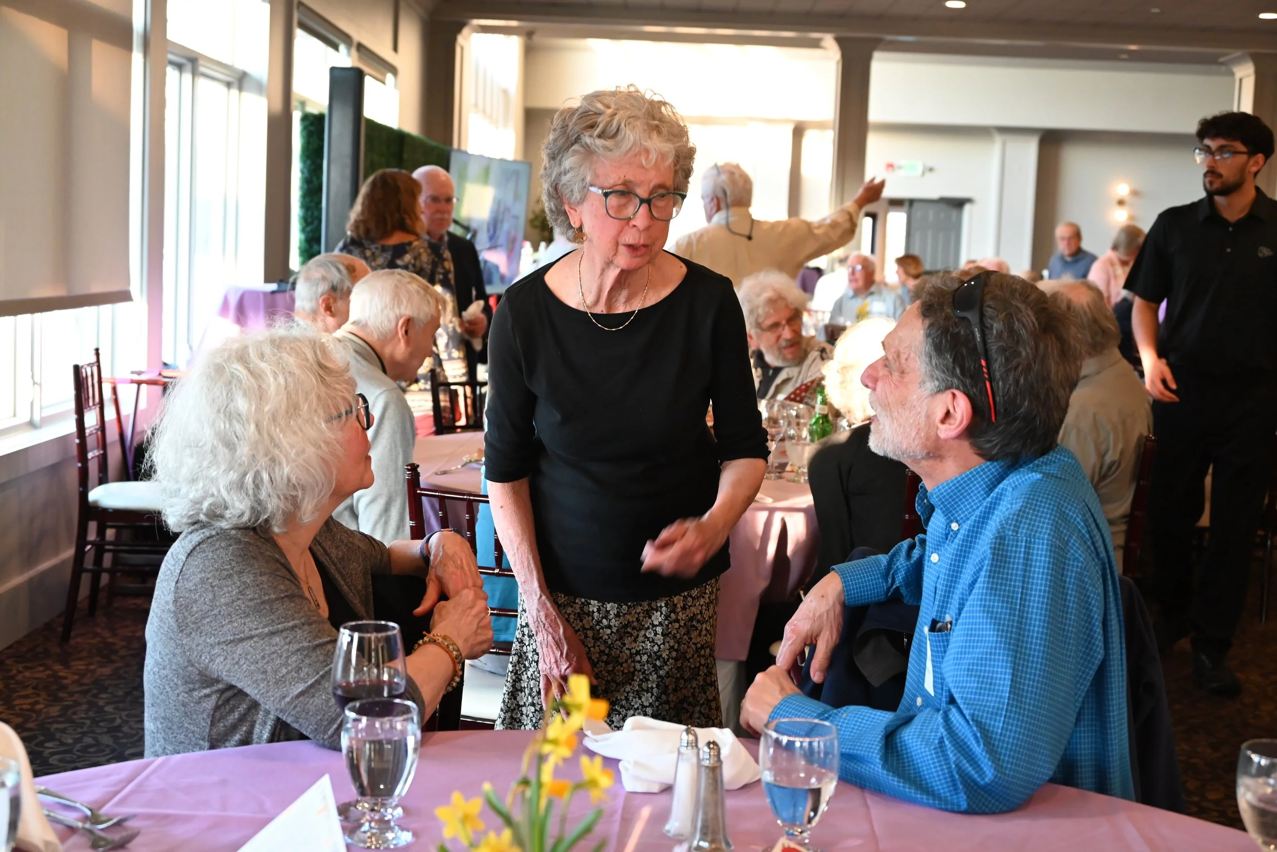 A group of elderly people at a restaurant, with a woman standing and greeting a seated man. Other guests are in the background, some talking and others looking around, with a waiter also visible.
