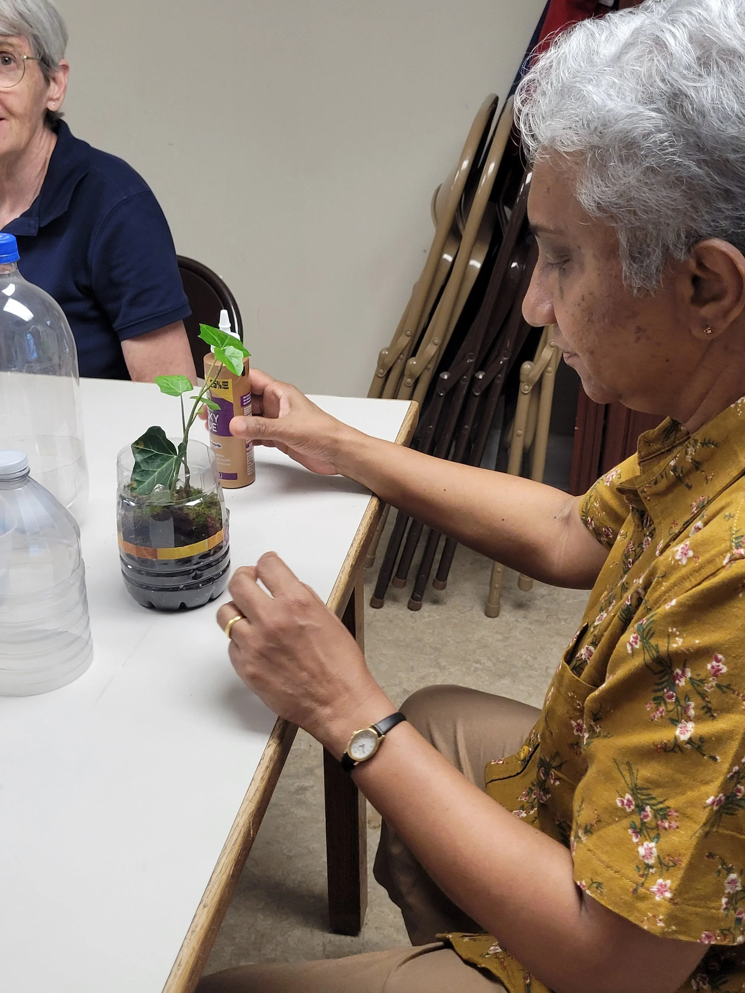 Older woman with short gray hair and earrings, wearing a watch and a yellow floral shirt, sitting at a table, holding a bottle with an artificial plant inside, with stacked chairs against the wall behind her.
