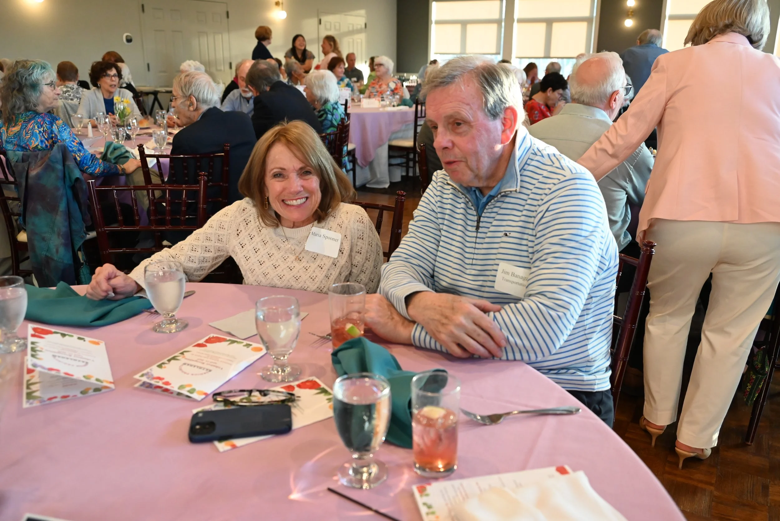 A woman smiling at a man at a social gathering, seated at a table with a pink tablecloth, drinks, and menus, while other people converse and eat in the background.