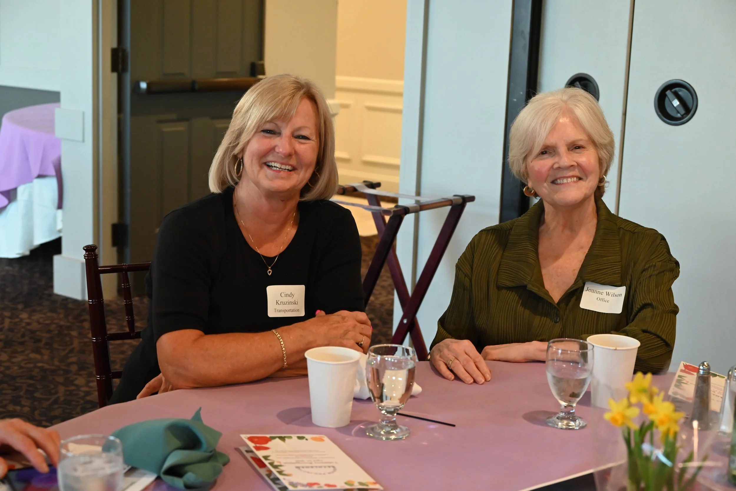 Two women sitting at a round table during a gathering or event, smiling at the camera, with name tags and drinks on the table.