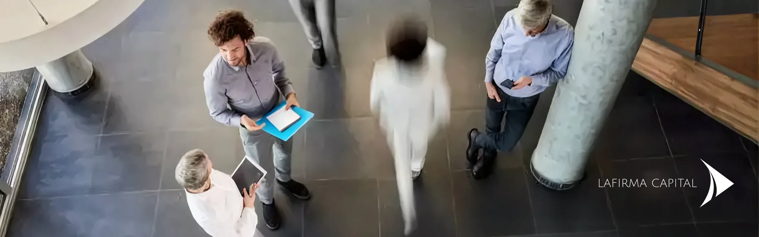 Imagen vista desde arriba de cinco personas en una oficina moderna, conversando y usando dispositivos electrónicos, con una columna grande y un área con piso de baldosas negras.