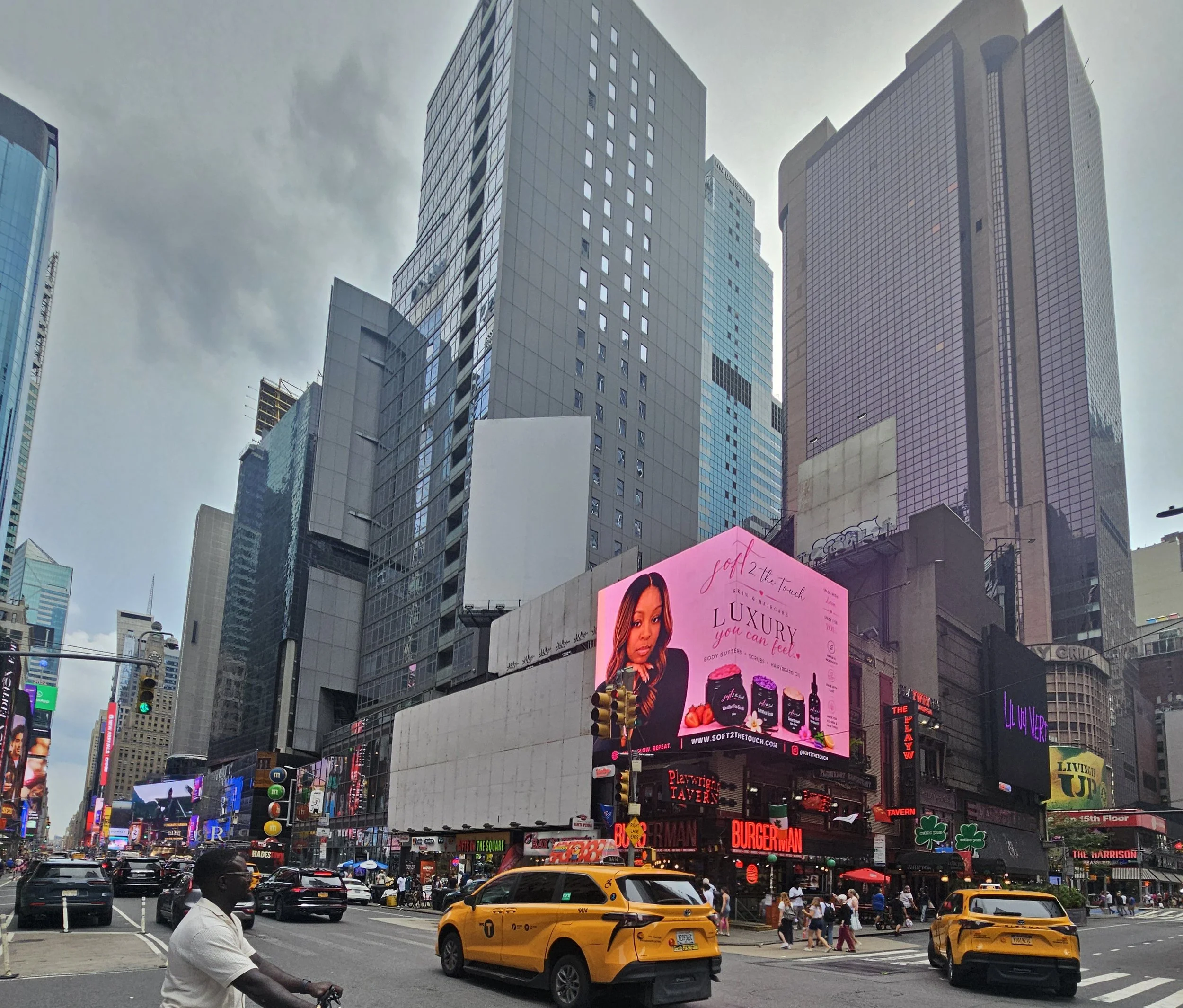 View of Times Square in New York City with tall buildings, digital billboards, and busy traffic. People walking, taxis, and numerous advertisements are visible.