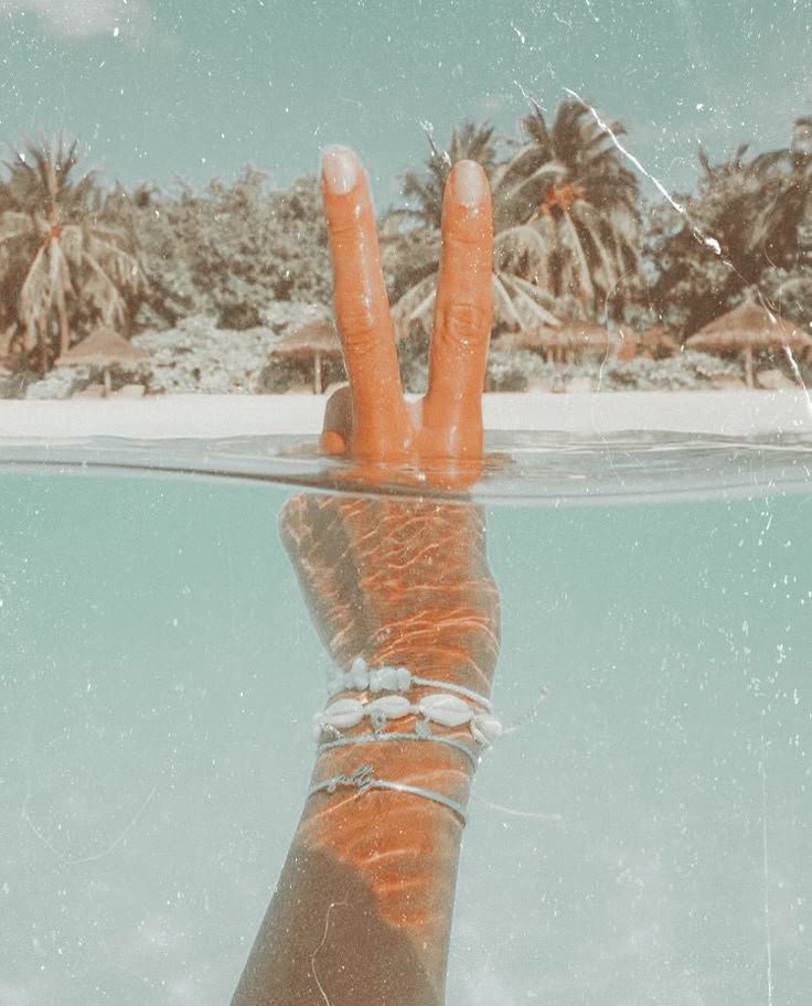 A hand making a peace sign partially submerged in a swimming pool with a tropical beach scene in the background.