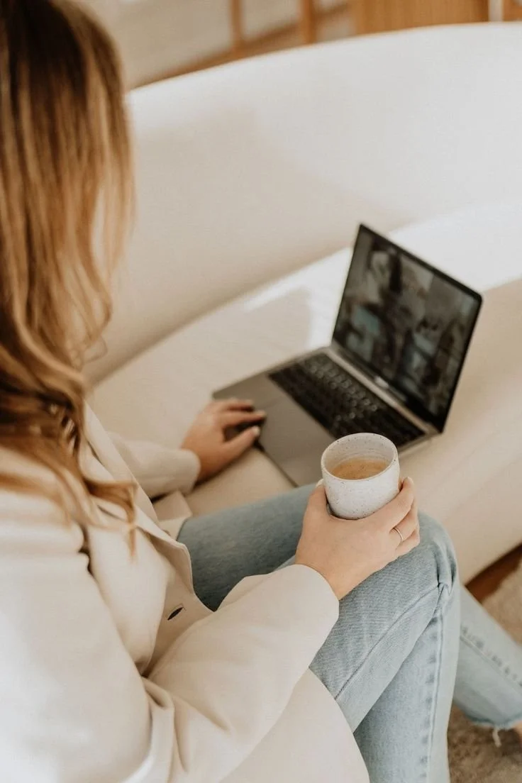 Person sitting on a light-colored couch holding a coffee mug while looking at an open laptop.