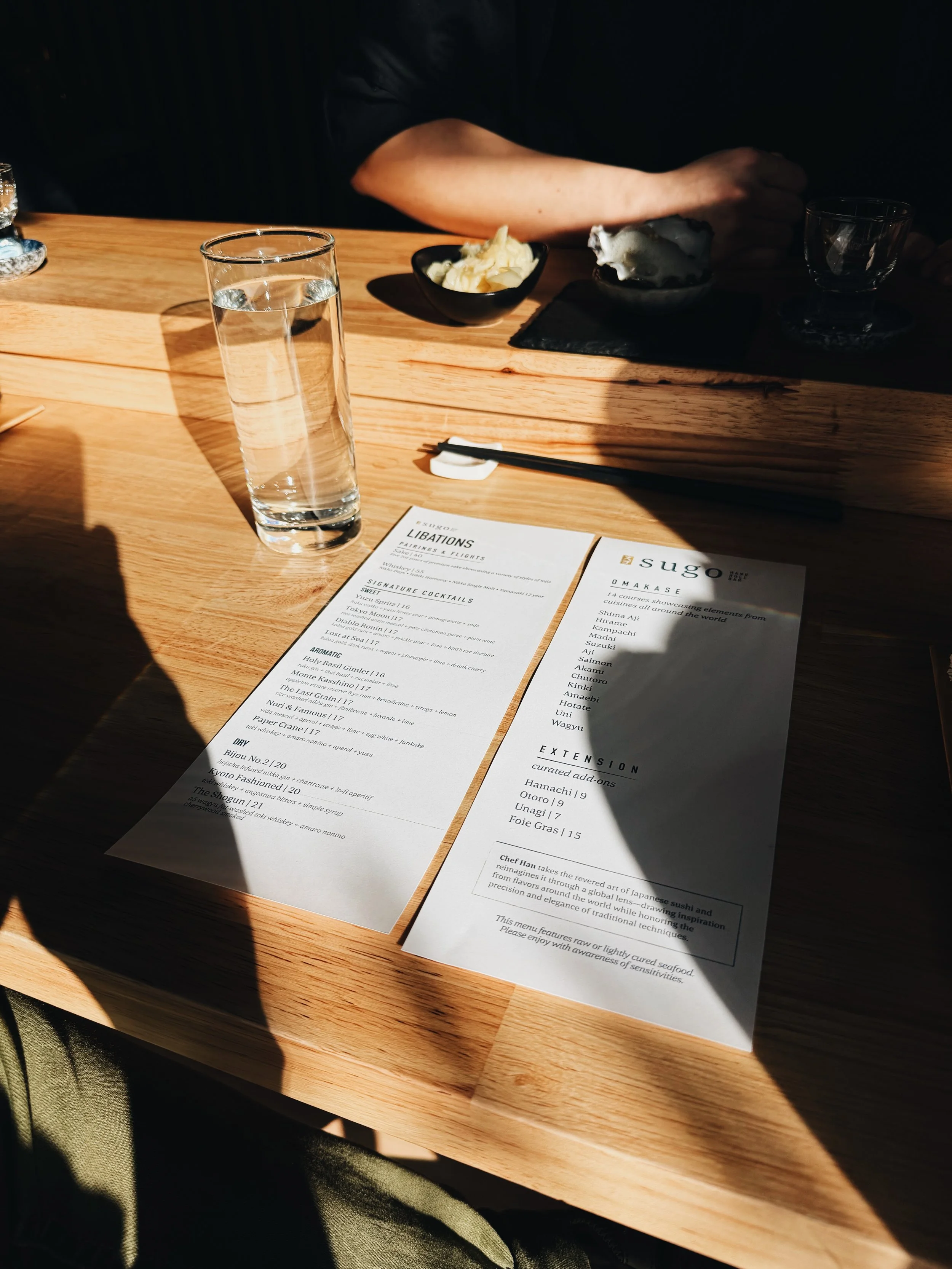 A wooden table with a tall glass of water, two menus, and dishes with butter and mackerel. Shadows cast across the table, and a person’s arm is visible in the background.