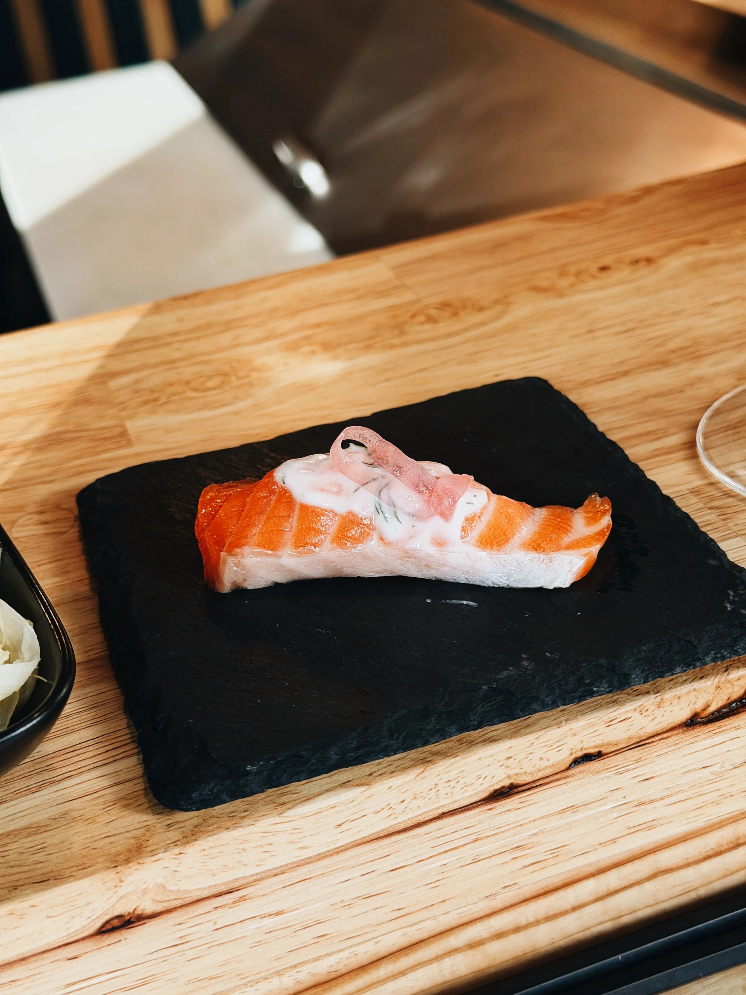 A piece of salmon sashimi topped with a small amount of white sauce, served on a black slate plate on a wooden surface.