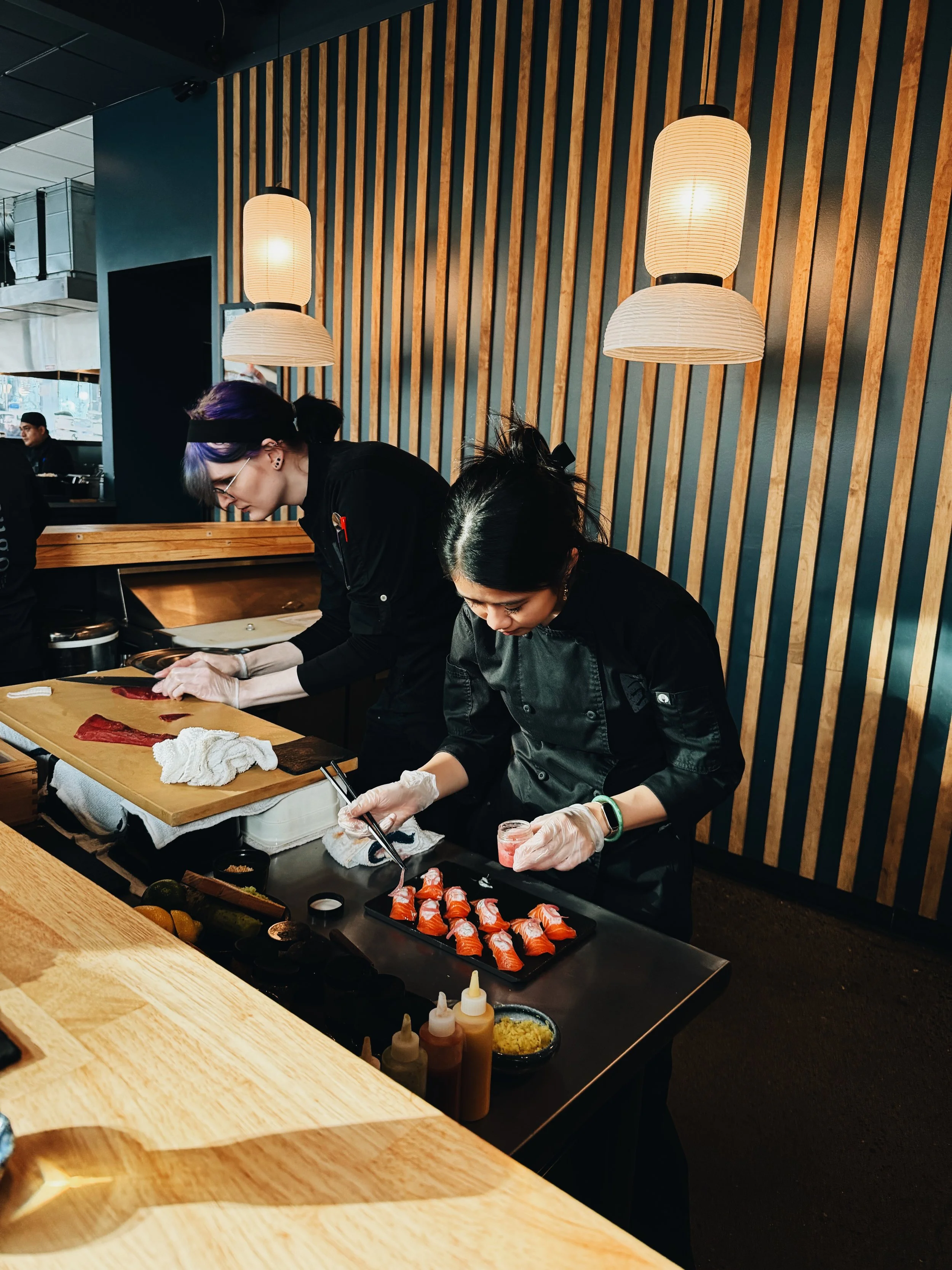 Two women working behind a sushi counter, preparing sushi rolls with sushi ingredients and utensils displayed on the black counter.