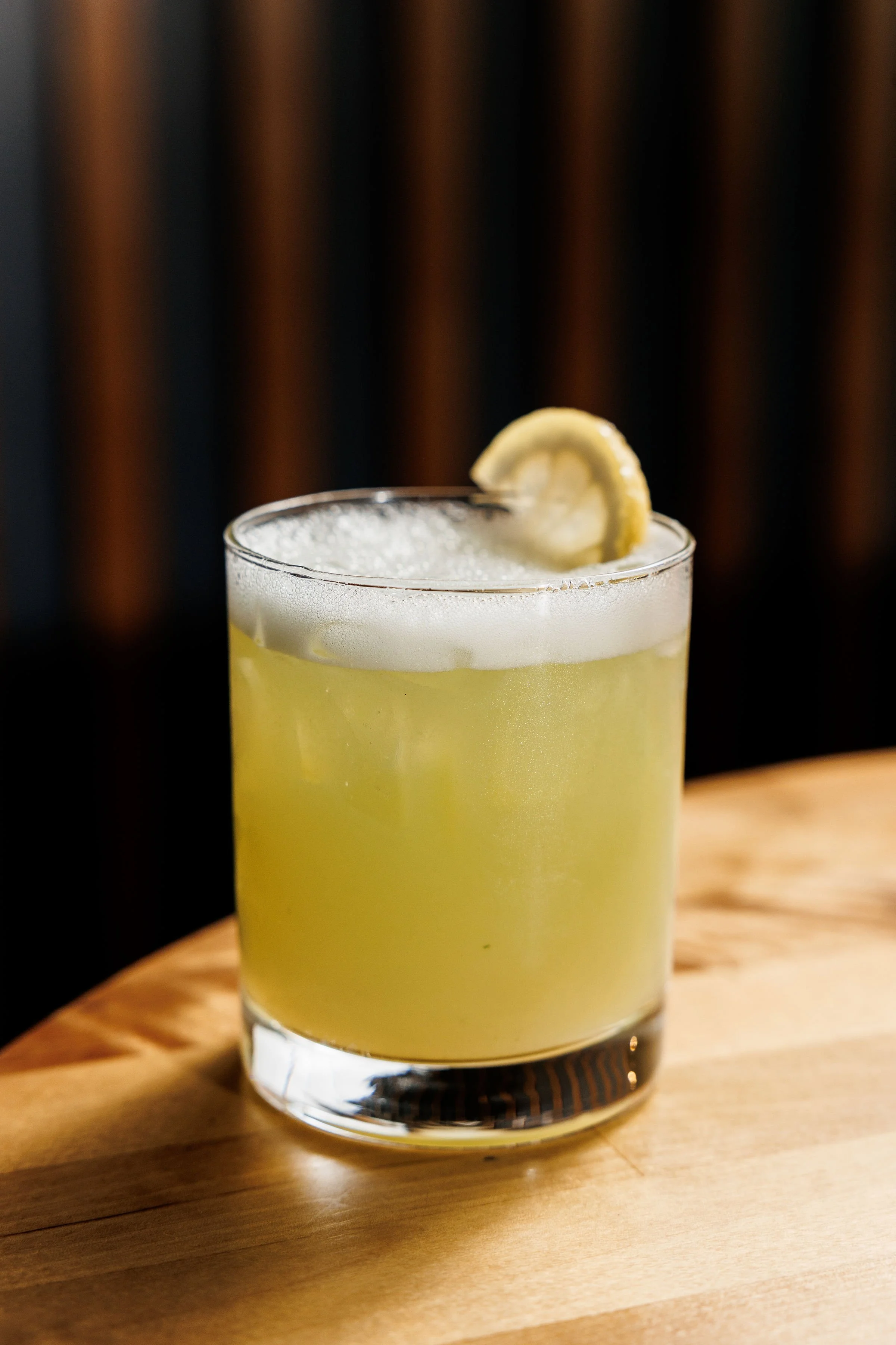 A cocktail in a glass garnished with a lemon wedge, sitting on a wooden surface with a dark background.