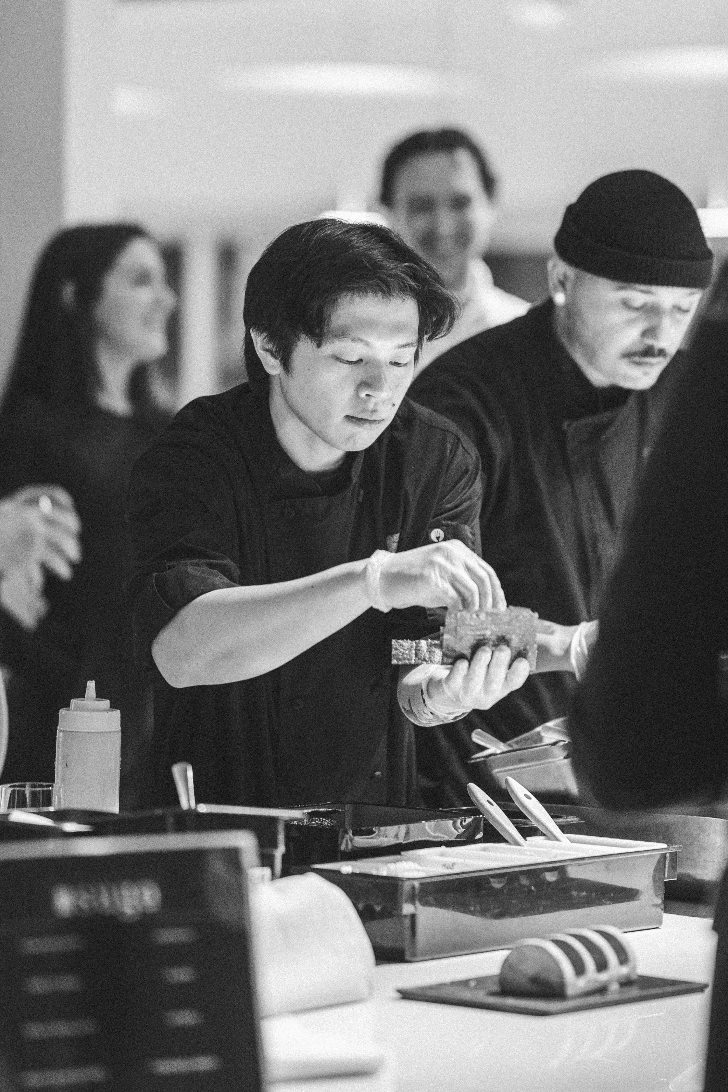People preparing food in a kitchen or restaurant, with the main focus on a woman wearing gloves and a man wearing a beanie, both working at a counter with various kitchen tools and ingredients.