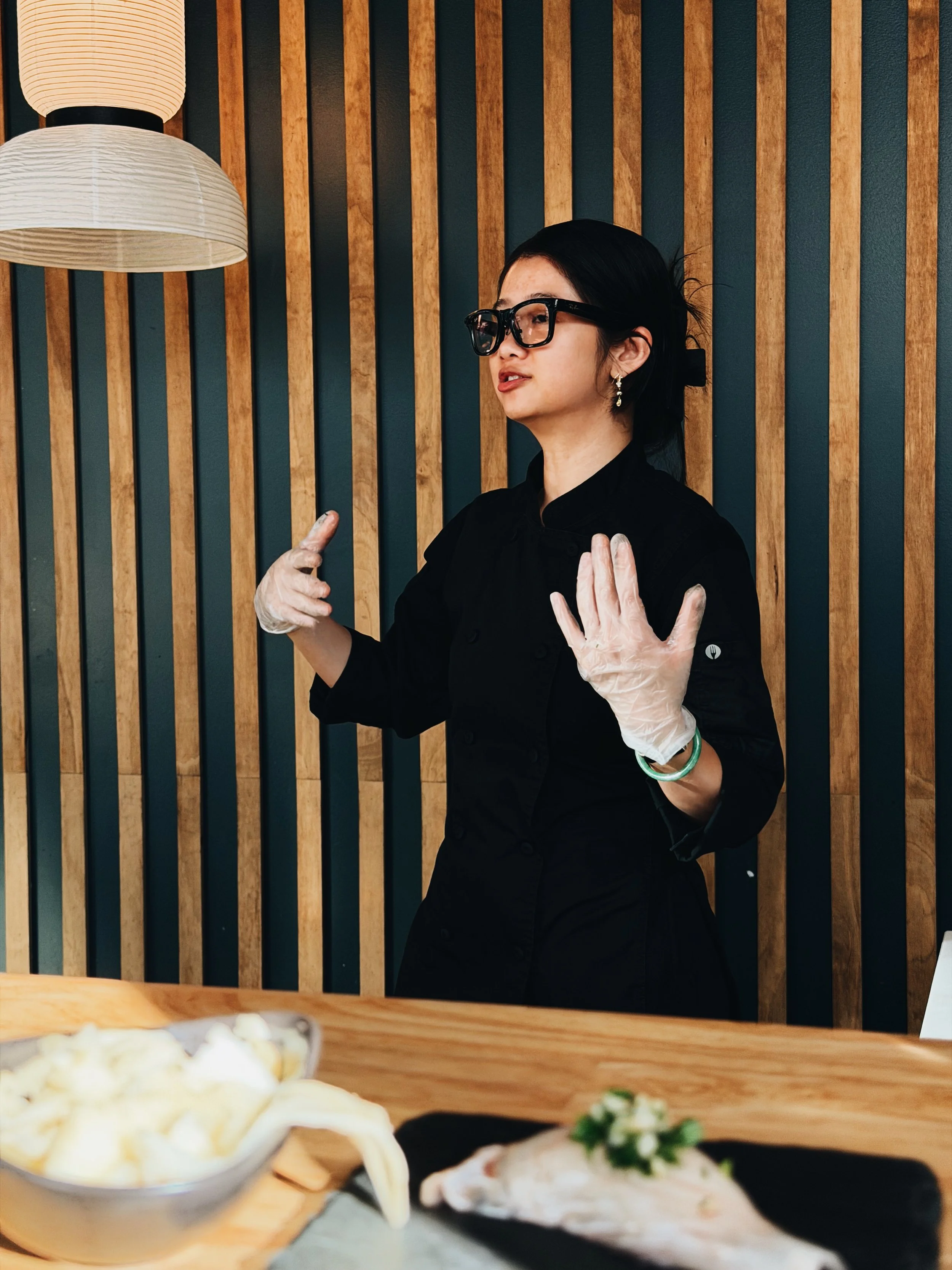 A woman in black clothes and glasses, wearing earrings, standing in front of a wooden wall, with her hands raised. Only the top of food dishes are visible at the bottom of the image.