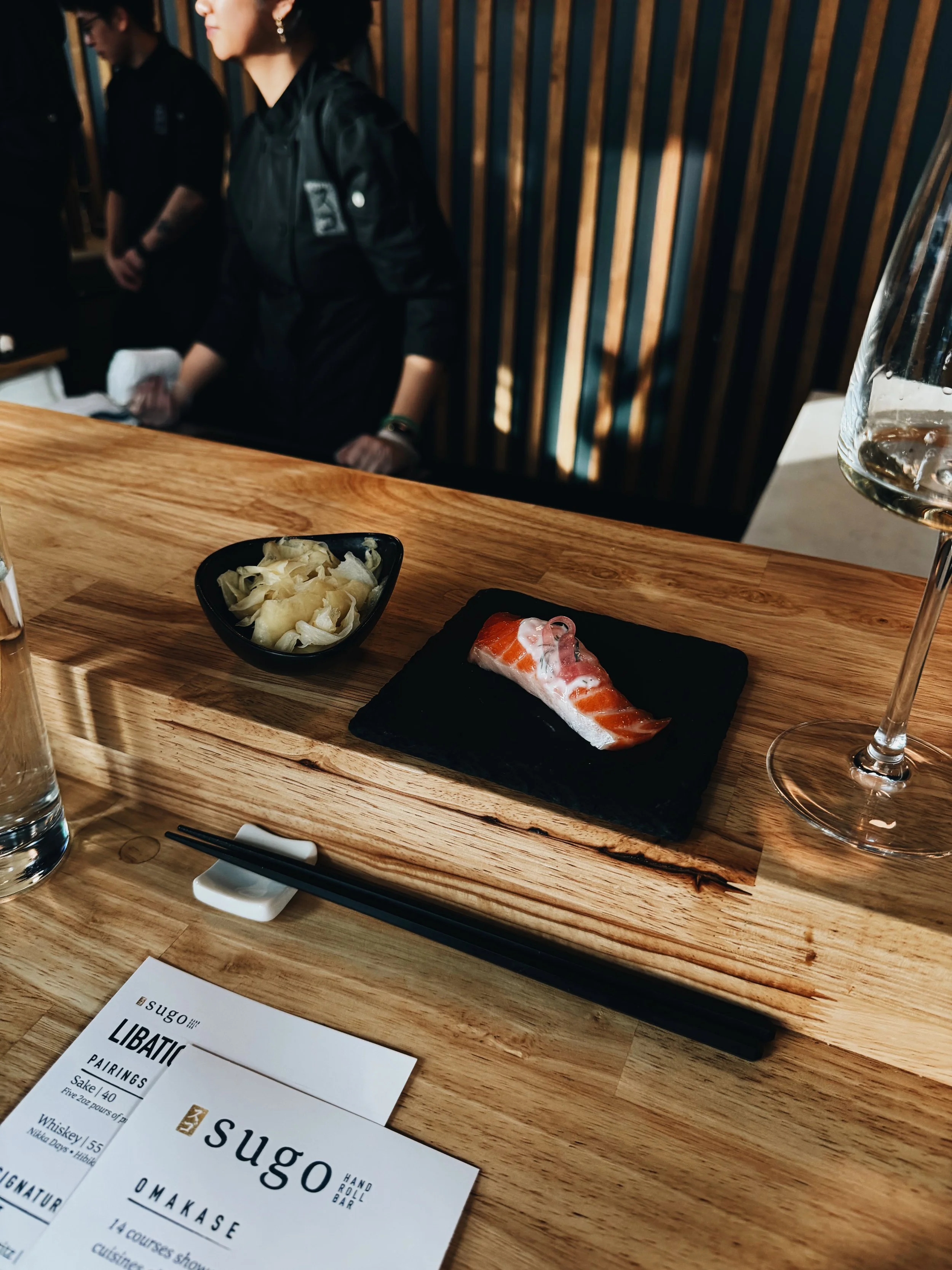 A wooden table set with a small black plate of pickled ginger, a piece of salmon sashimi on a black tray, a glass of water and a wine glass. In the background, two people in black clothing and earrings, with a wooden slat wall behind them. A menu she