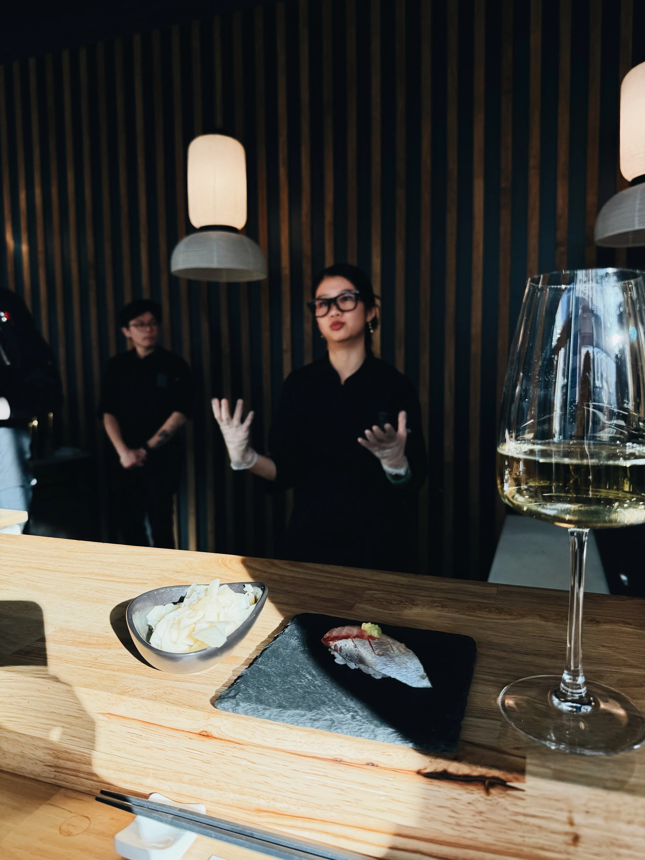 Close-up of a wooden table with a bowl of sliced creamy cheese, a black slate plate with a piece of sushi topped with wasabi, a glass of white wine, and a woman in black glasses and clothing gesturing with her hands, with two other women in black in 