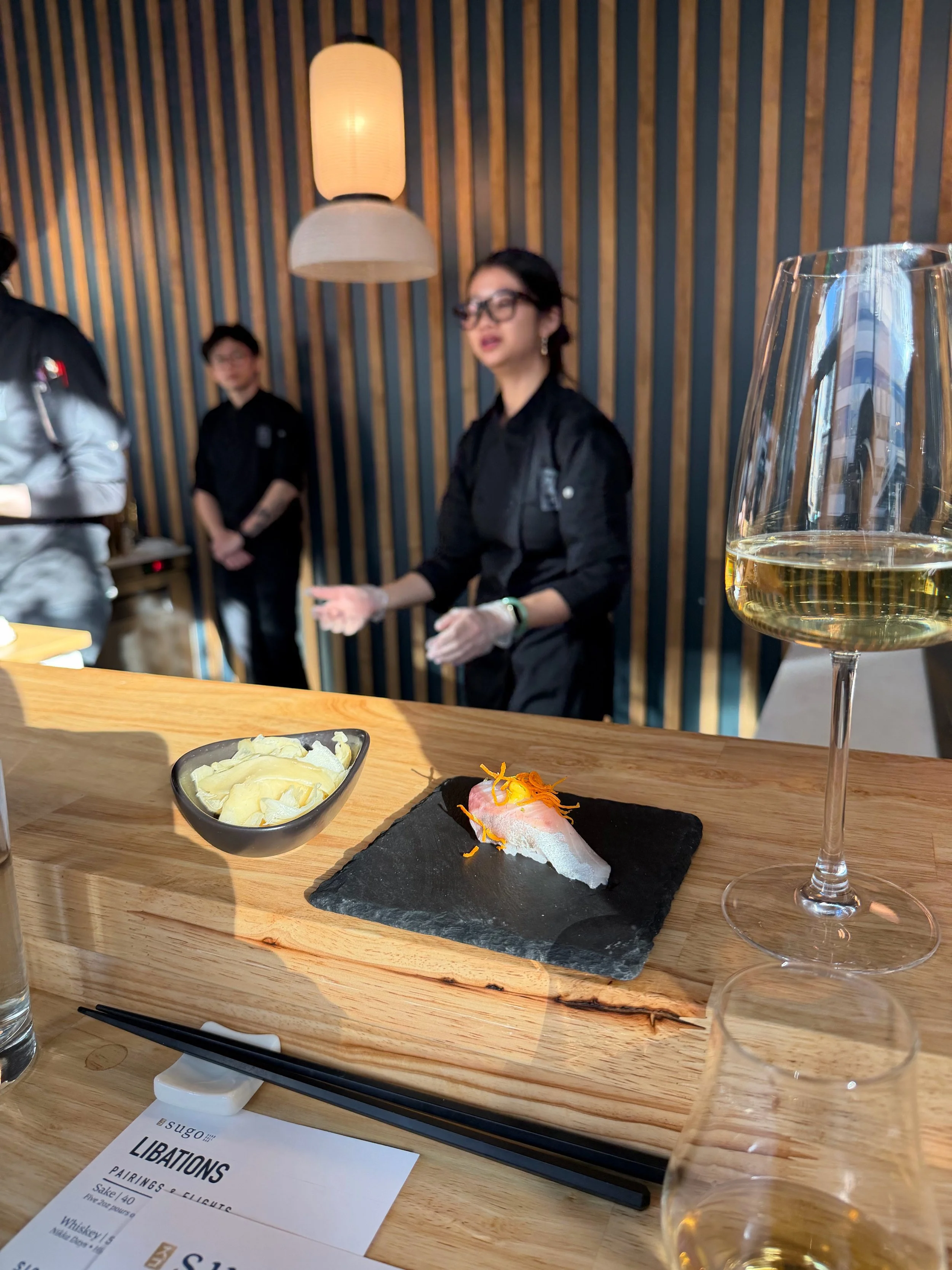 A sushi roll on a black stone serving board with shredded carrots on top, a small bowl of potato chips, a glass of white wine, and a pair of black chopsticks on a wooden table at a restaurant with people in the background.