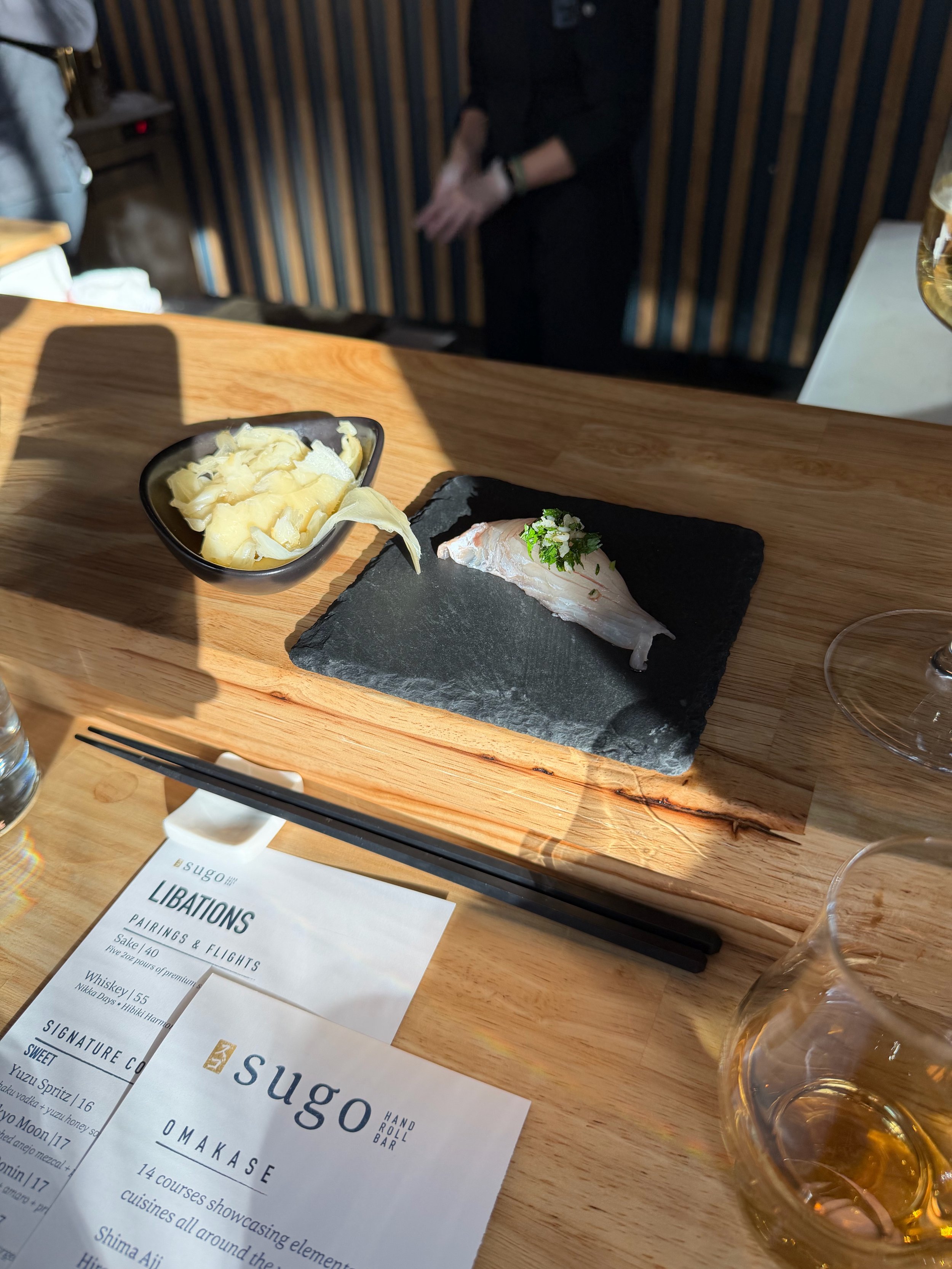 A wooden table with a black slate plate holding a piece of fish garnished with herbs, a black bowl filled with cheese, a glass of whiskey, and a menu from Sugo restaurant.