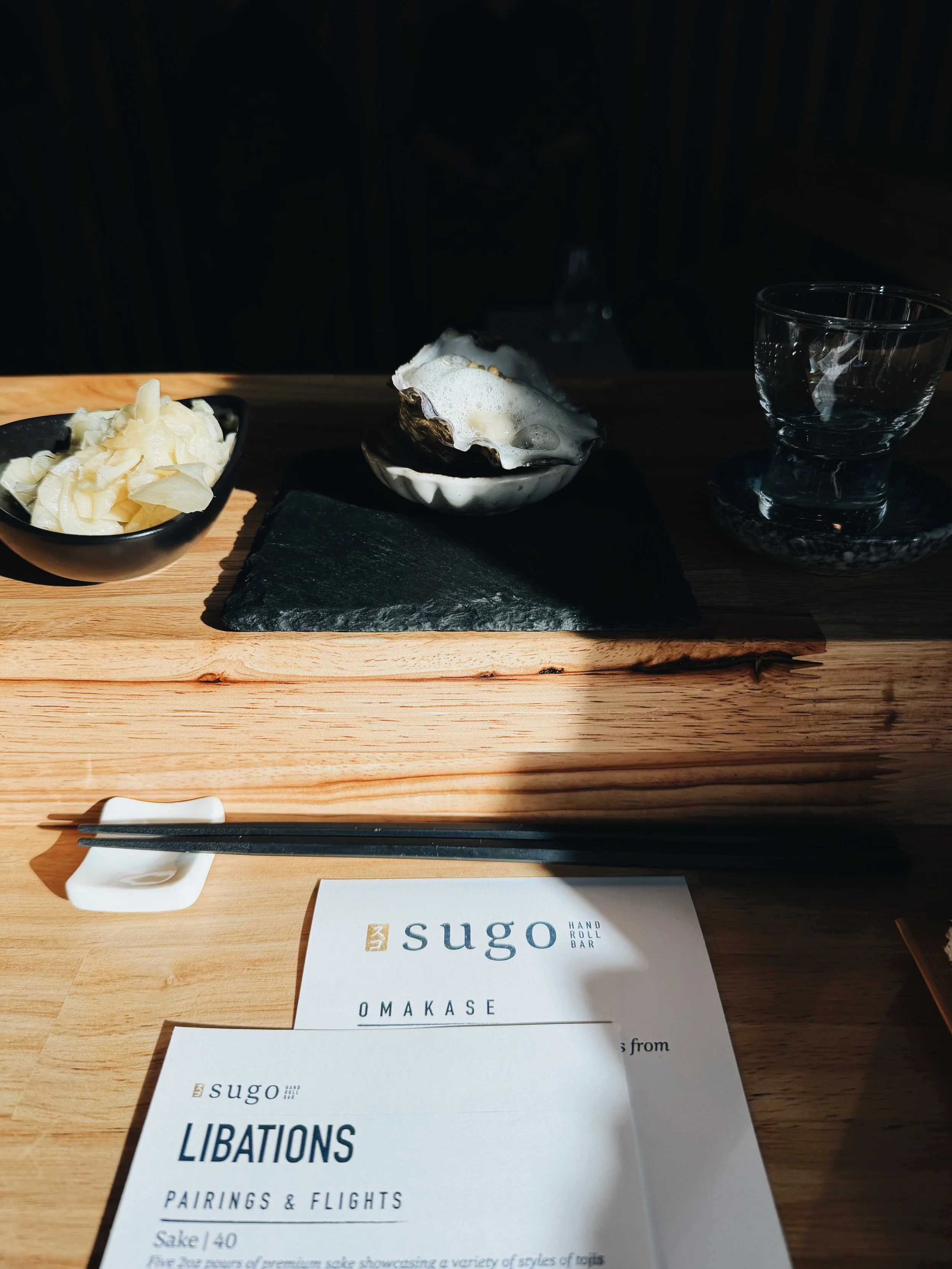 A wooden table with a black bowl of sliced white vegetables, a white scallop shell with foam, an empty glass bowl, black chopsticks on a white chopstick rest, and printed menus and name cards for Sugo Hand Roll Bar.