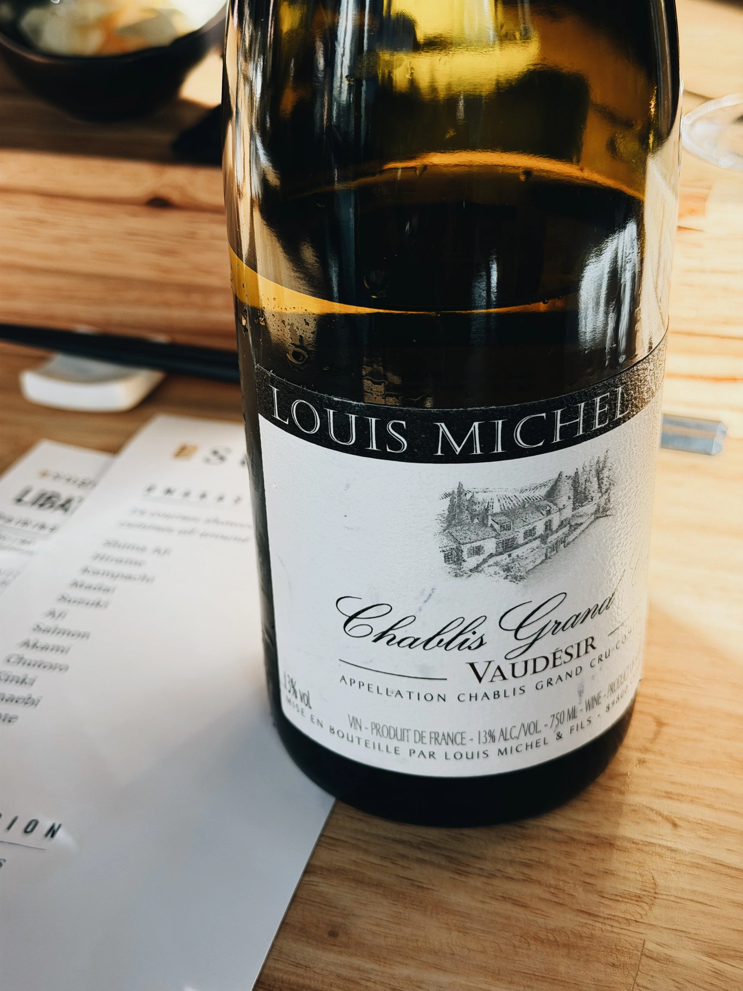 A wine bottle labeled Louis Michel Chablis Grand Vaudesir with a detailed black and white illustration of a vineyard or farm landscape, placed on a wooden table with brochures or menus nearby.