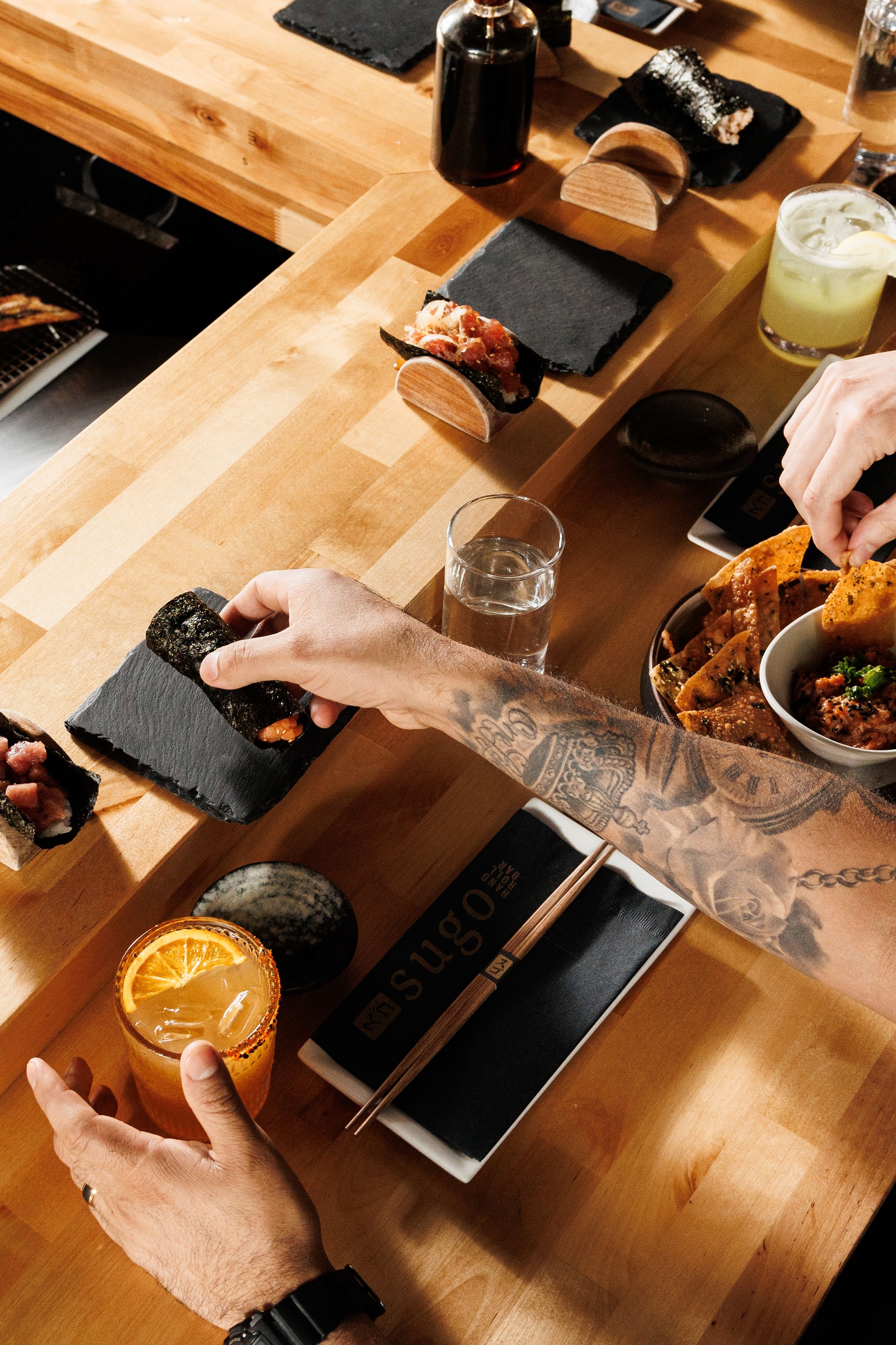 An overhead view of a wooden table with Japanese sushi rolls, a glass of orange-colored drink, and several drinks. Two people are reaching for food tips, and a tattooed arm is visible holding a sushi roll.