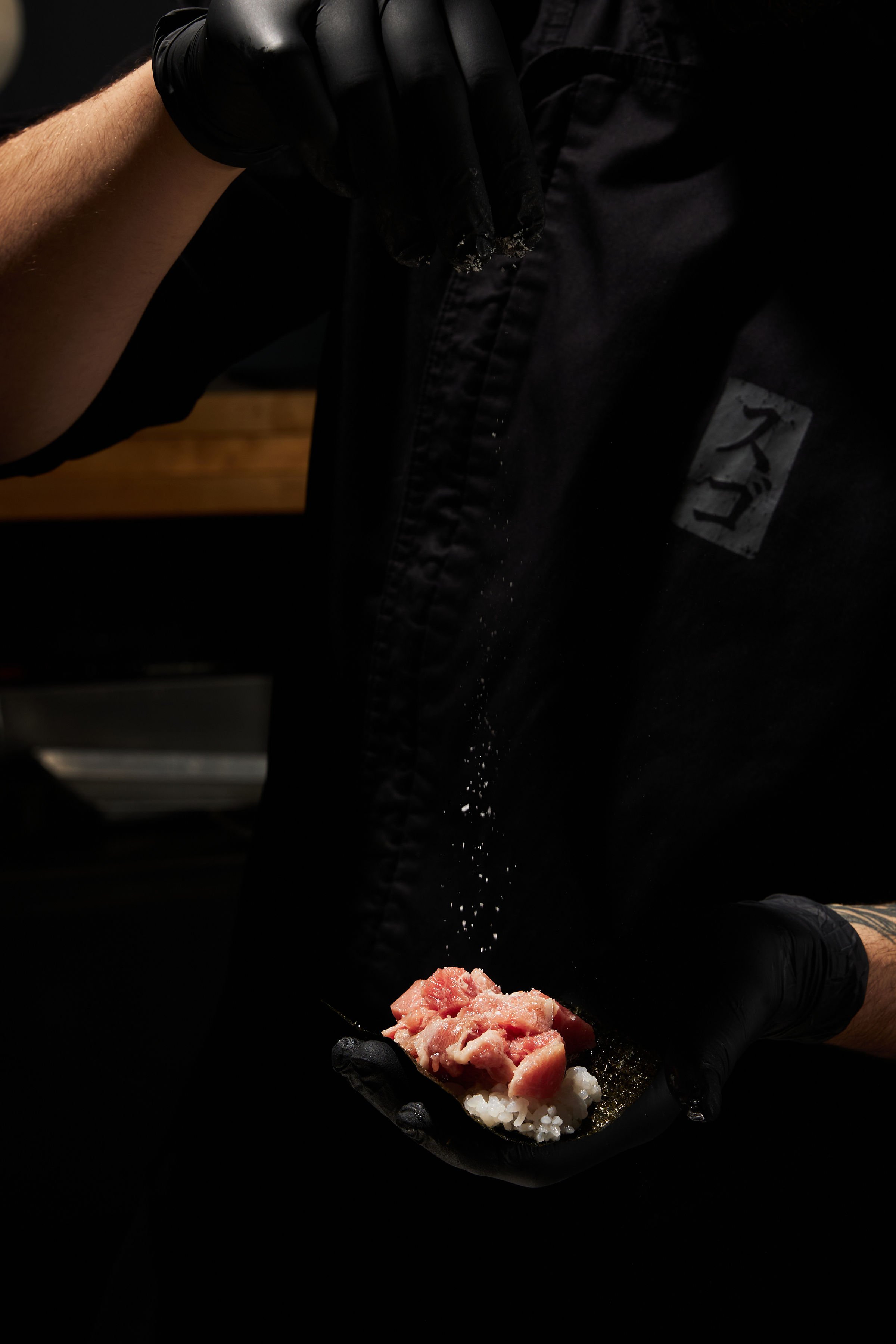 Person wearing black gloves preparing sushi, holding a piece of raw fish over a bed of rice on a nori seaweed sheet.