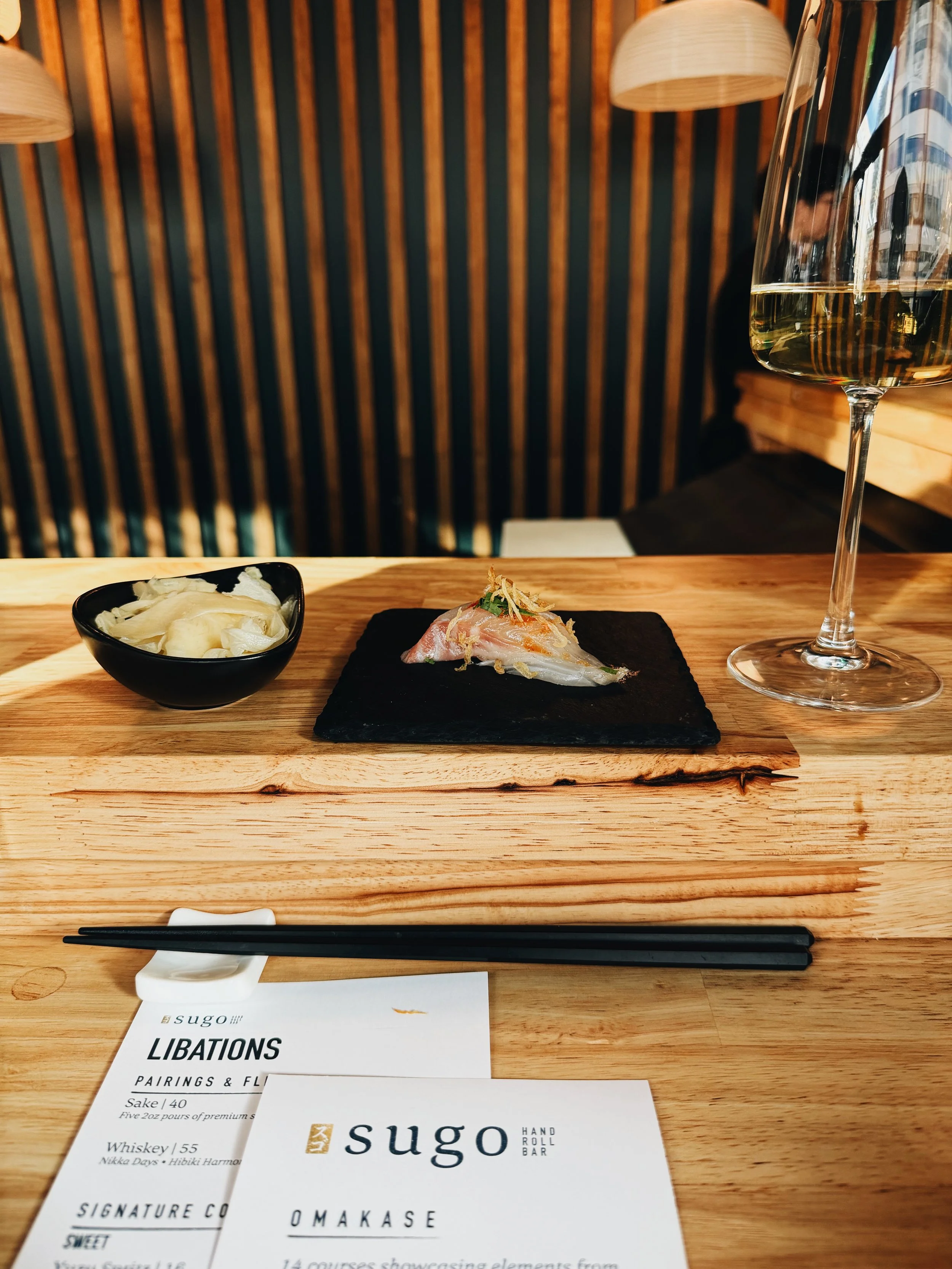 A wooden table in a restaurant with a small dish of pickled ginger, a slate plate with a slice of sashimi garnished with fried shallots, a glass of white wine, chopsticks on a napkin, and a menu from Sugo Hand Roll Bar.