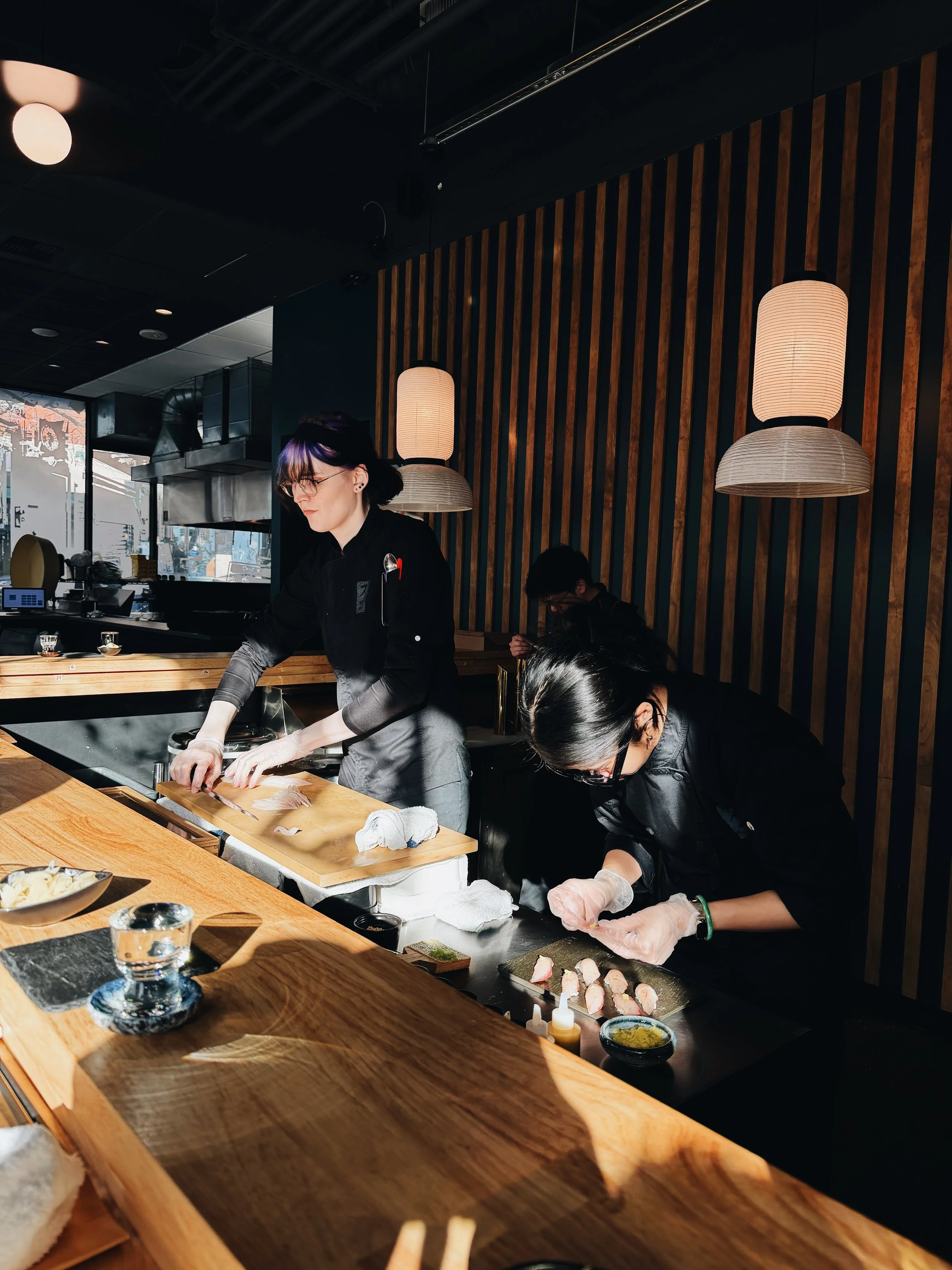 People preparing sushi at a restaurant counter with decorative hanging lamps and wood-paneled wall.