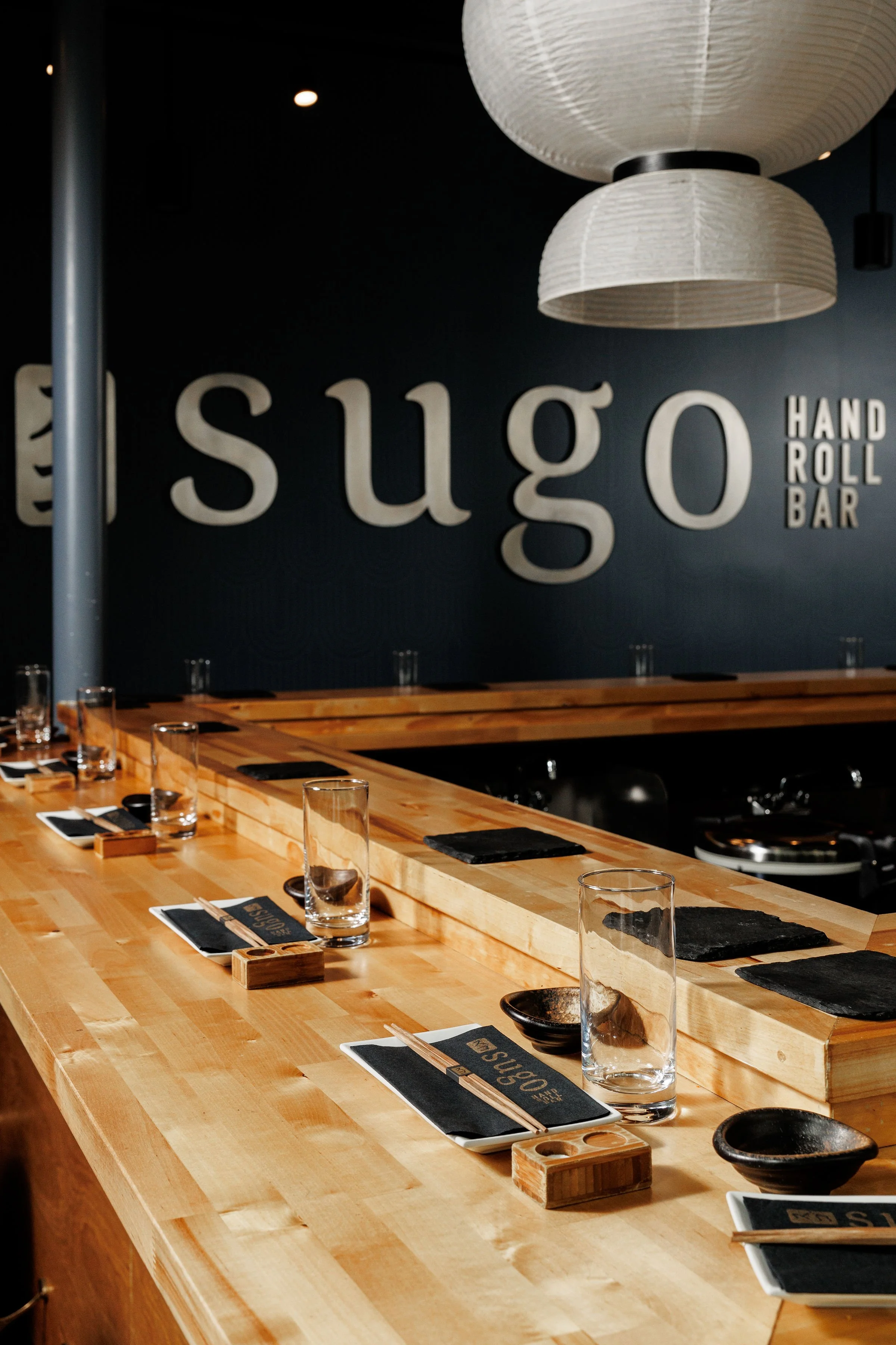 Interior of a Japanese sushi bar with a wooden counter set with glasses, small plates, chopsticks, and black cloth napkins, with a dark wall in the background that has the word 'sugo' and 'hand roll bar' written on it, and large hanging white paper lanterns overhead.