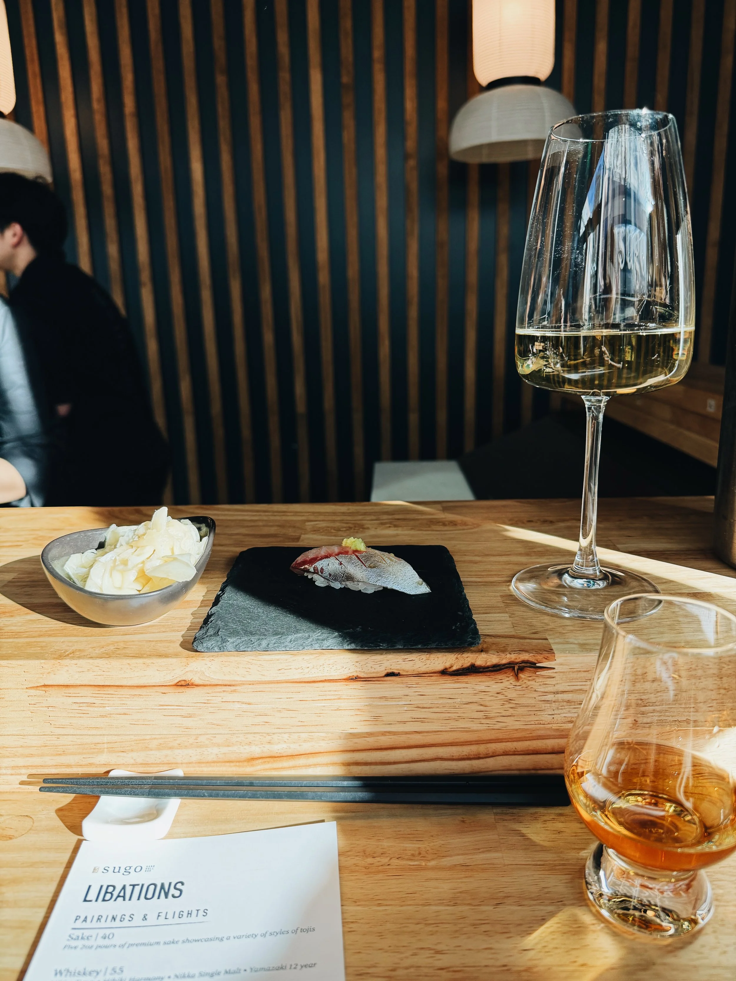A dining table with a glass of white wine, a small dish of whipped cream, a plate of sushi, and a menu. The background features a wooden wall with hanging lamps, and a person sitting to the left.