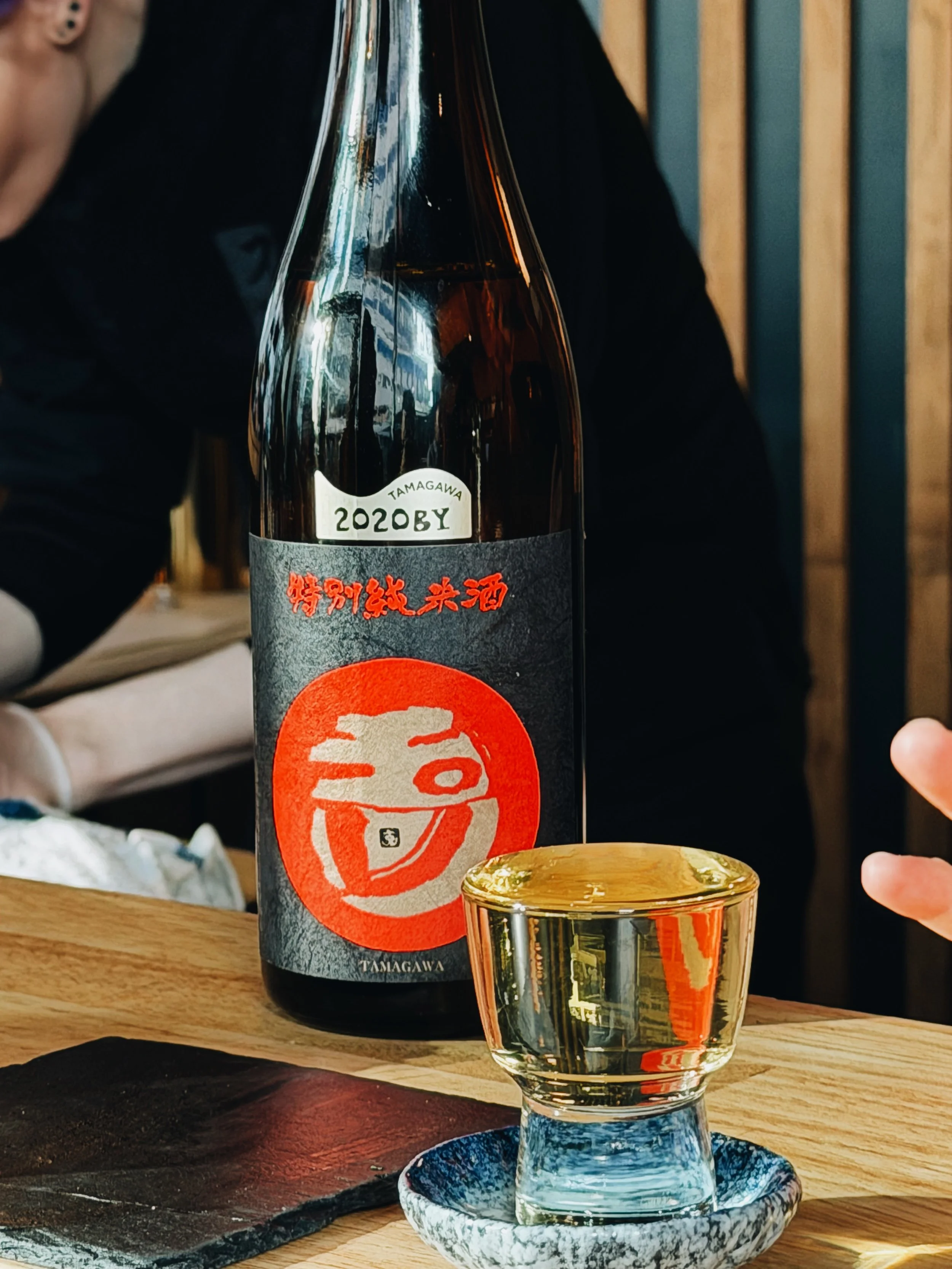 A glass of sake on a wooden table with a bottle of sake in the background. The glass is filled with a clear, pale yellow liquid and is placed on a small, blue and white ceramic plate.