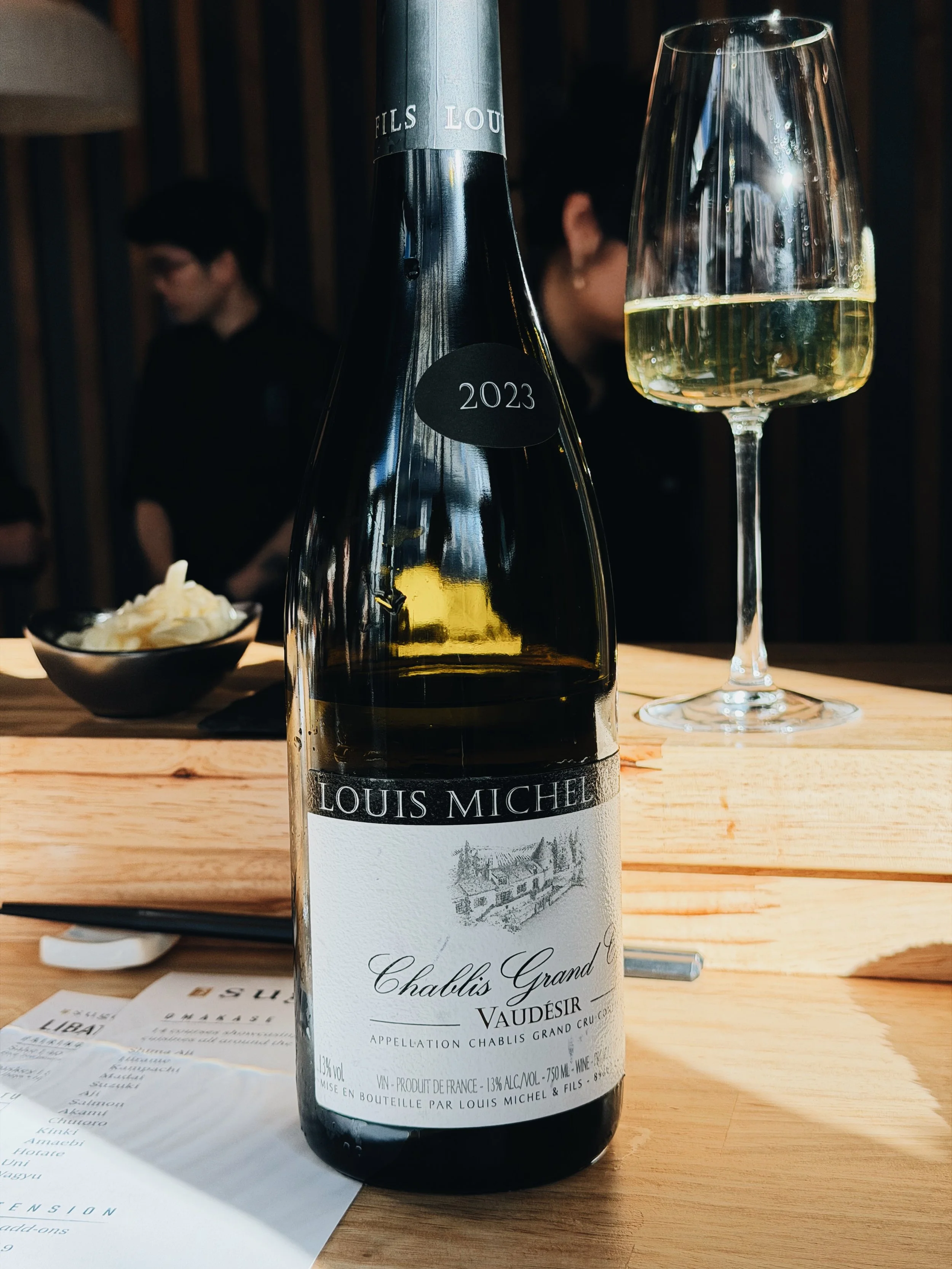Bottle of Louis Michel Chablis Grand Vaudésir white wine on a wooden table, with a glass of white wine beside it and a bowl of whipped cream in the background.