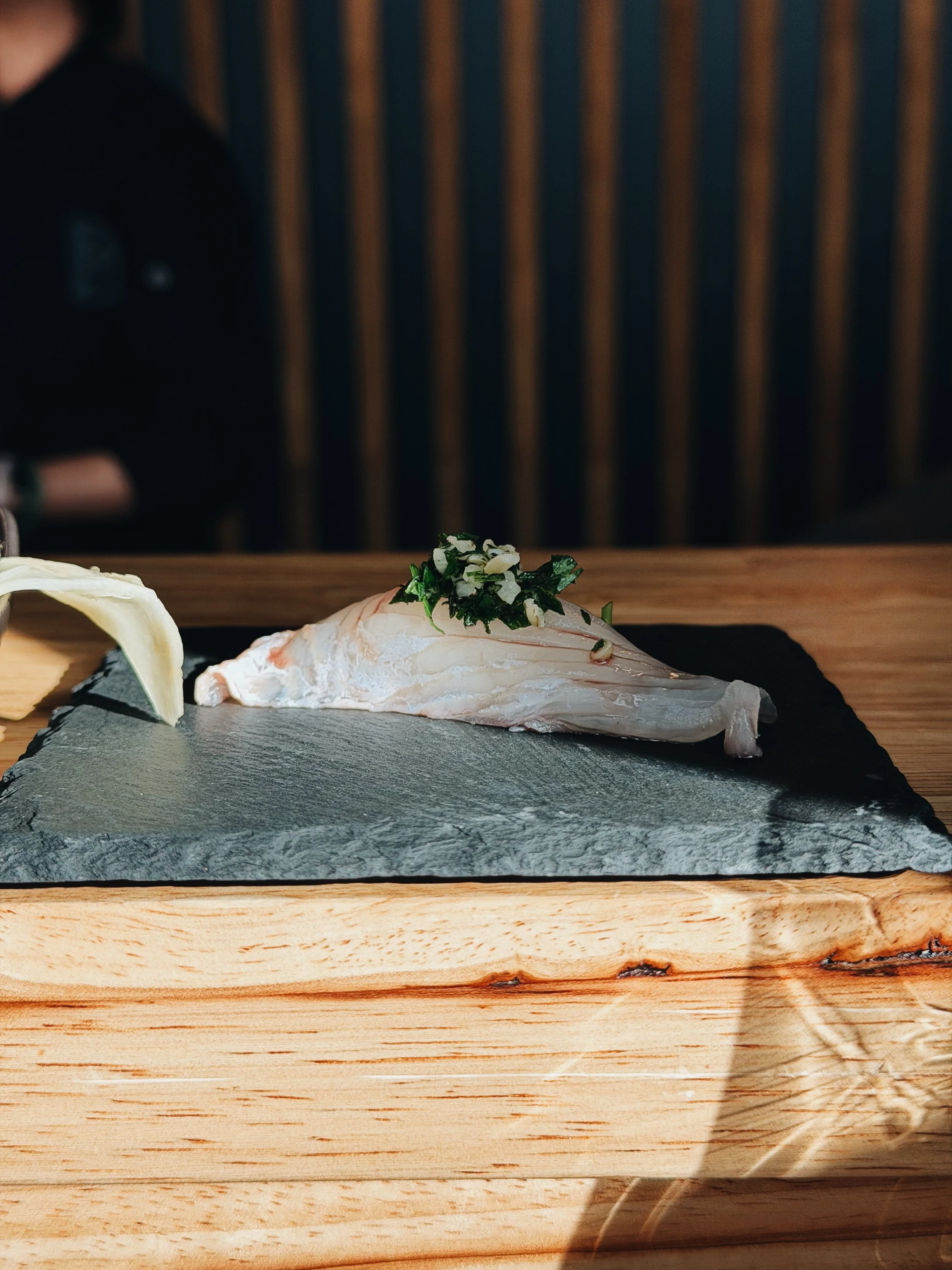 A slice of fish with herbs and garlic on top, placed on a black slate serving tray on a wooden table.