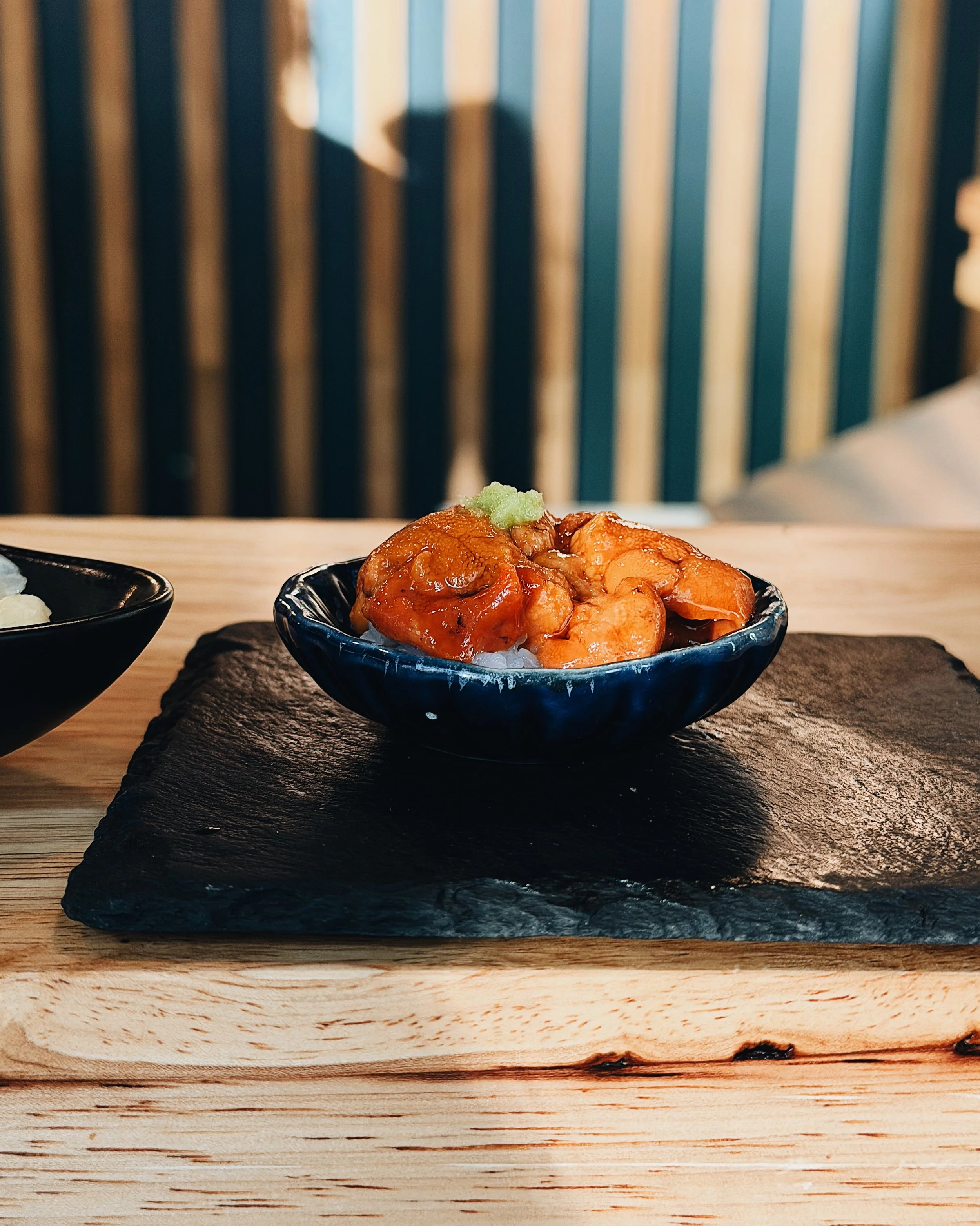 Close-up of a bowl of sashimi topped with wasabi, placed on a black slate platter on a wooden table, with a blurred background.