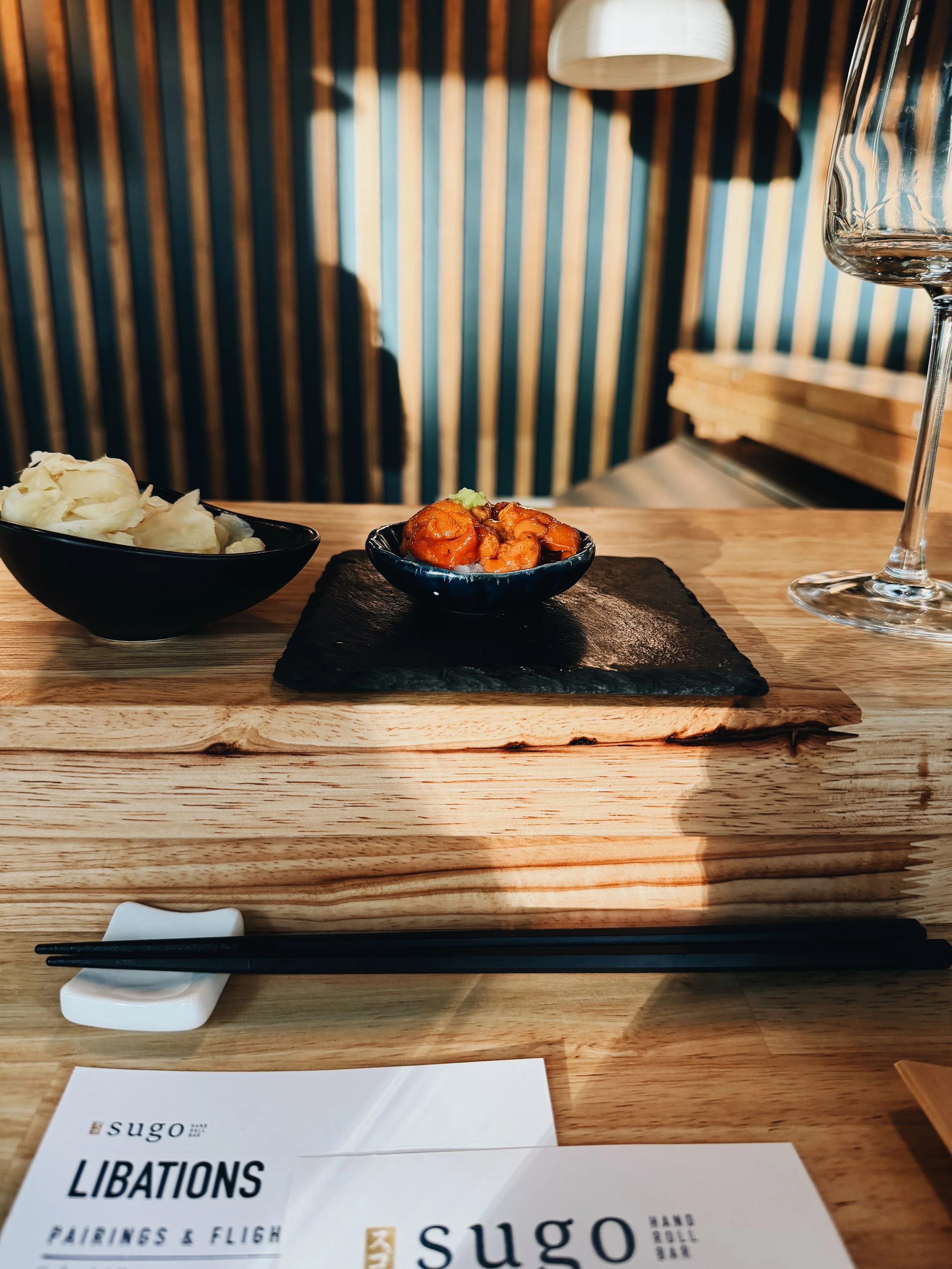 A wooden table set with black dishes containing pickled ginger and orange fish roe, a wine glass, black chopsticks on a white chopstick rest, and a menu or brochure with the text 'Sugo' and 'Libations'. In the background, there are striped wallpaper 