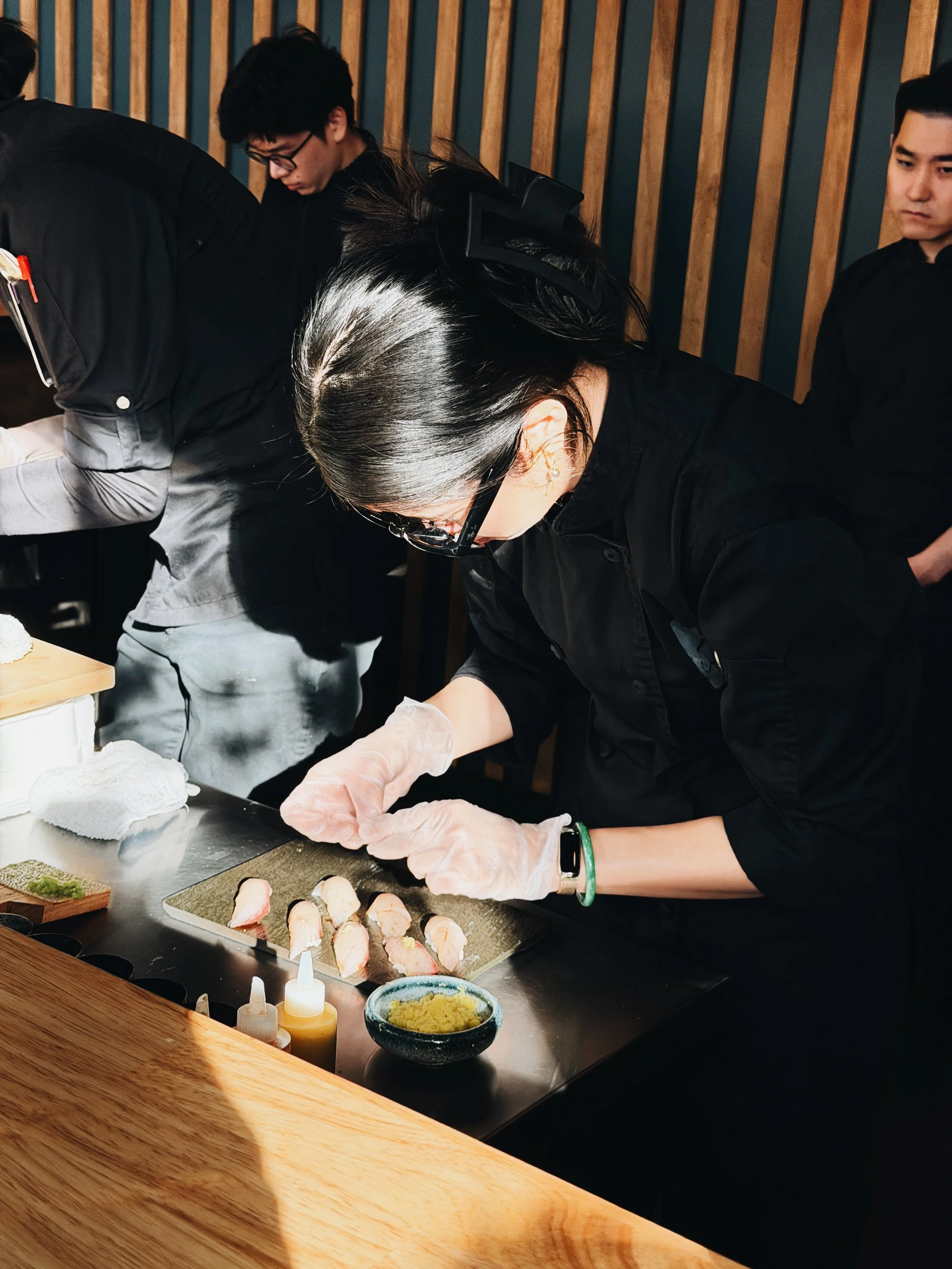 A person with black hair and glasses preparing sushi at a restaurant, surrounded by sushi rolls, sauces, and condiments on the counter.
