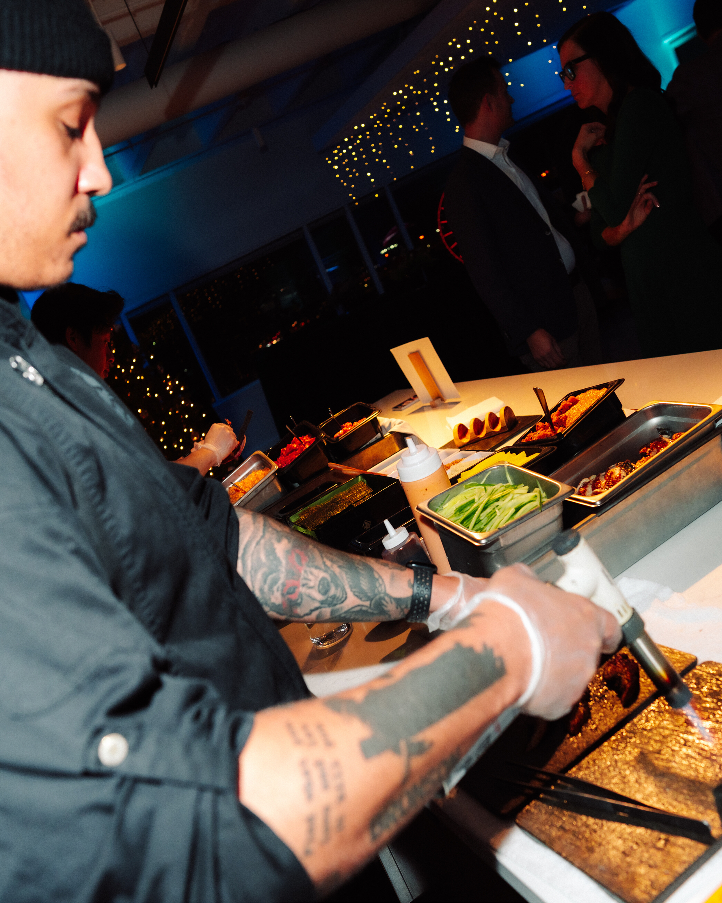 Person with tattoos on arm serving food at a buffet, with other people in the background in a dimly lit room with string lights on the ceiling.