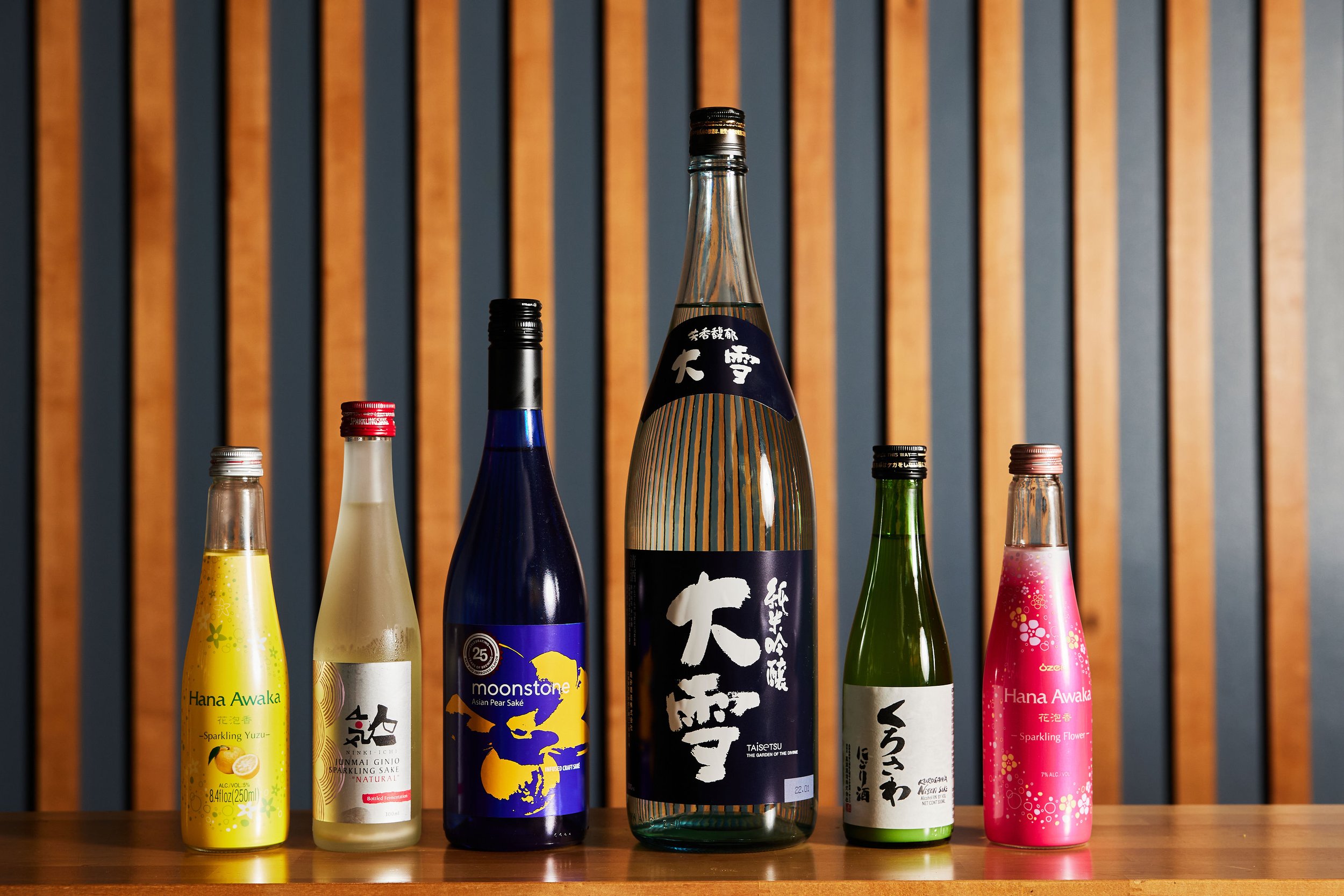 A collection of seven colorful bottles of sake and sparkling drinks on a wooden shelf with a wooden and gray striped wall in the background.
