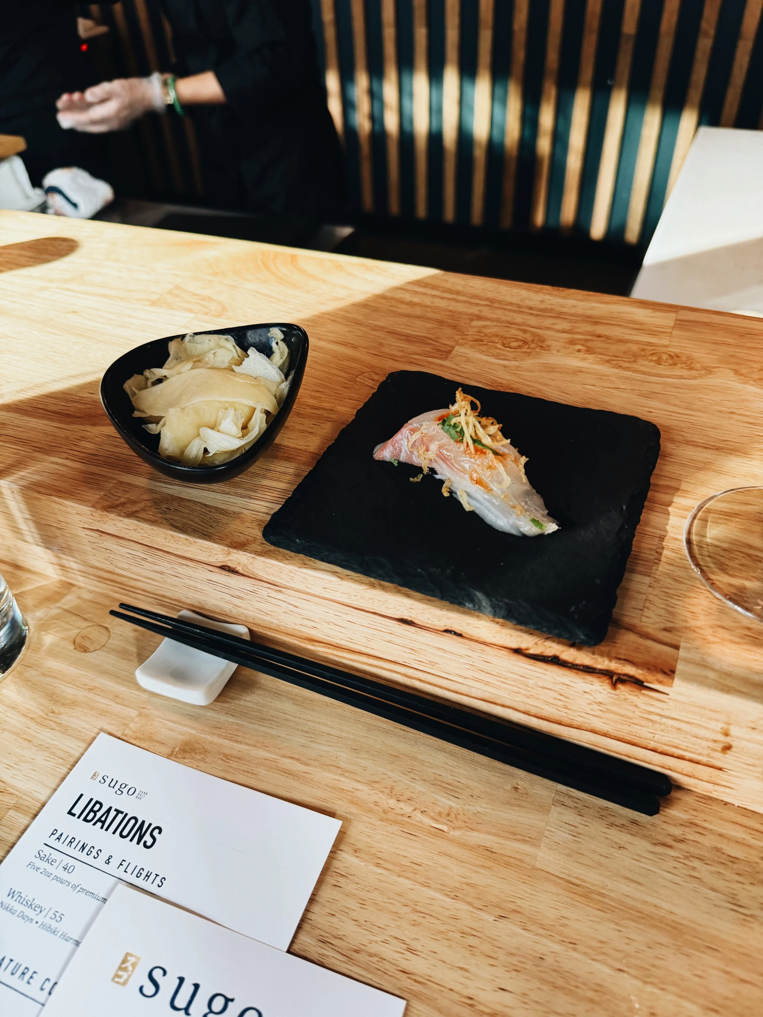 A wooden table with a black slate serving board holding a piece of fish garnished with herbs and fried shallots, a small black bowl of pickled ginger, a pair of black chopsticks, and a menu from Sugo on the table.