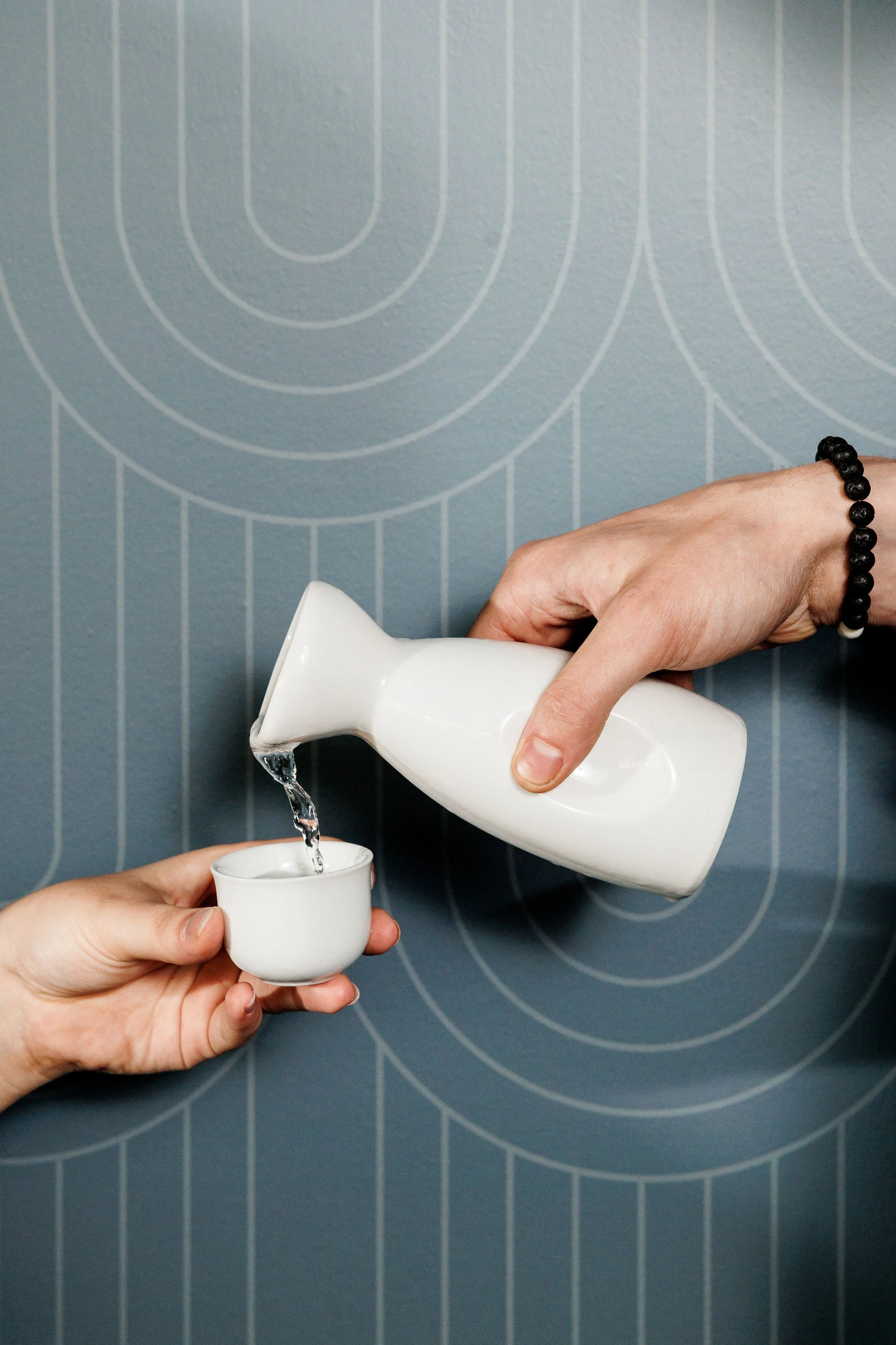 A person is pouring water from a white ceramic teapot into a small white cup held by another hand. The background is a blue wall with concentric circle patterns.