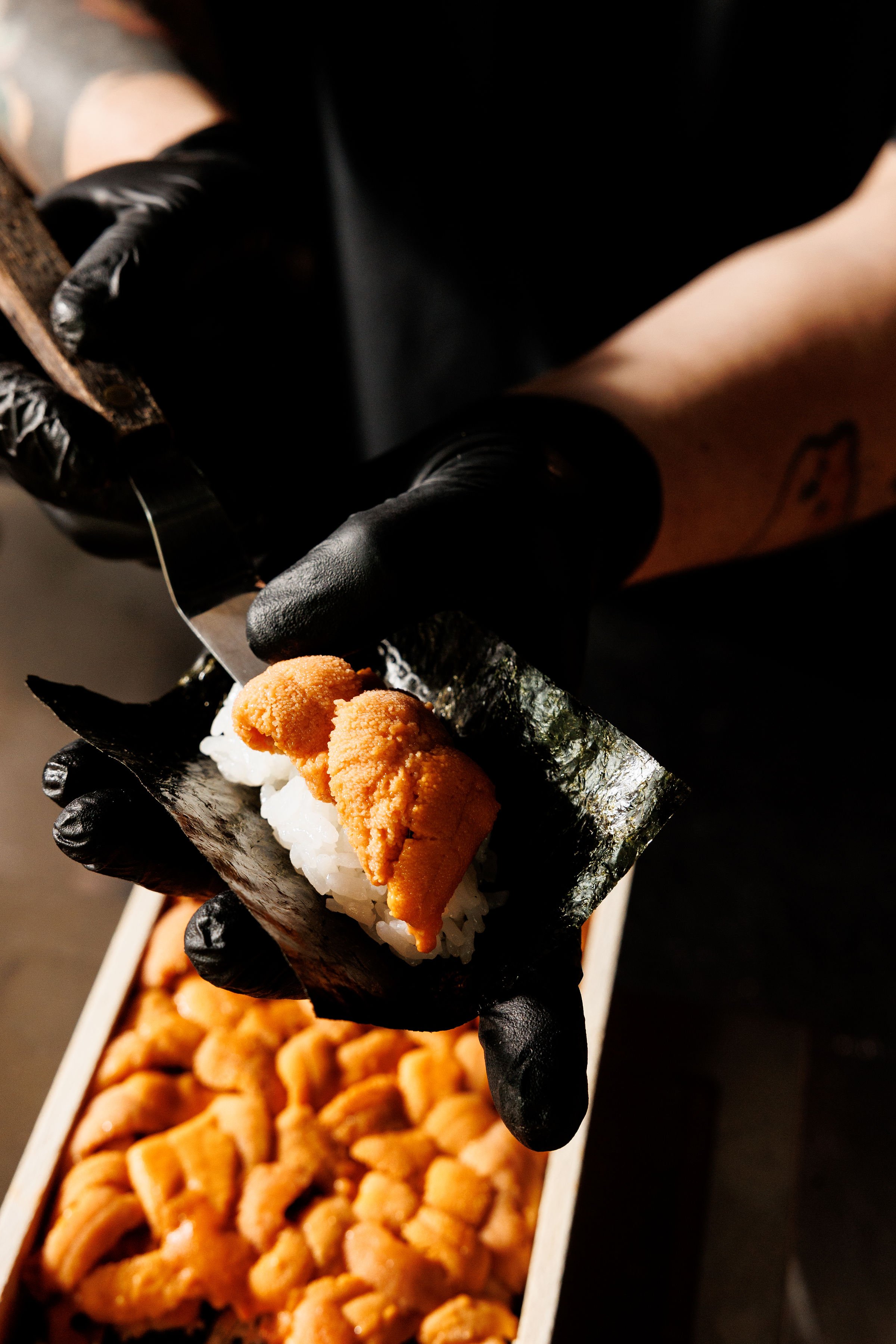 Close-up of a gloved hand holding a piece of sushi with sea urchin and rice. In the background, a tray with more sea urchin sushi.