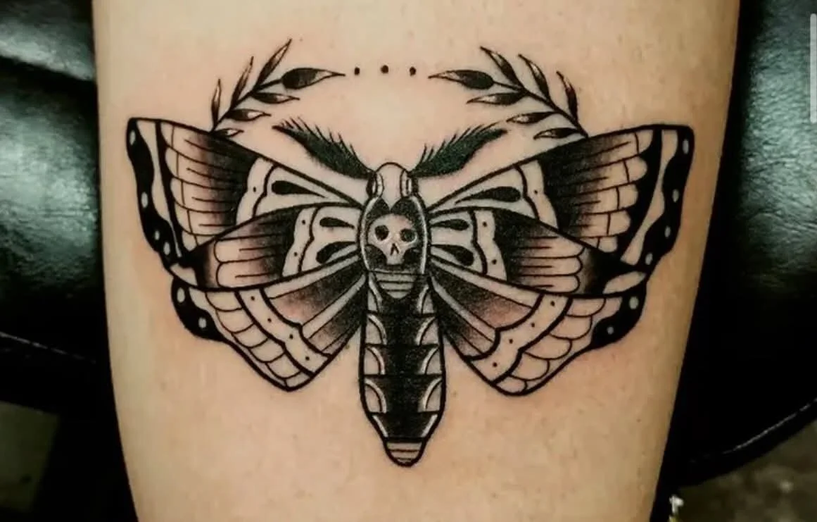 Tattoo of a moth with a skull in the center, surrounded by decorative lines and leaves.