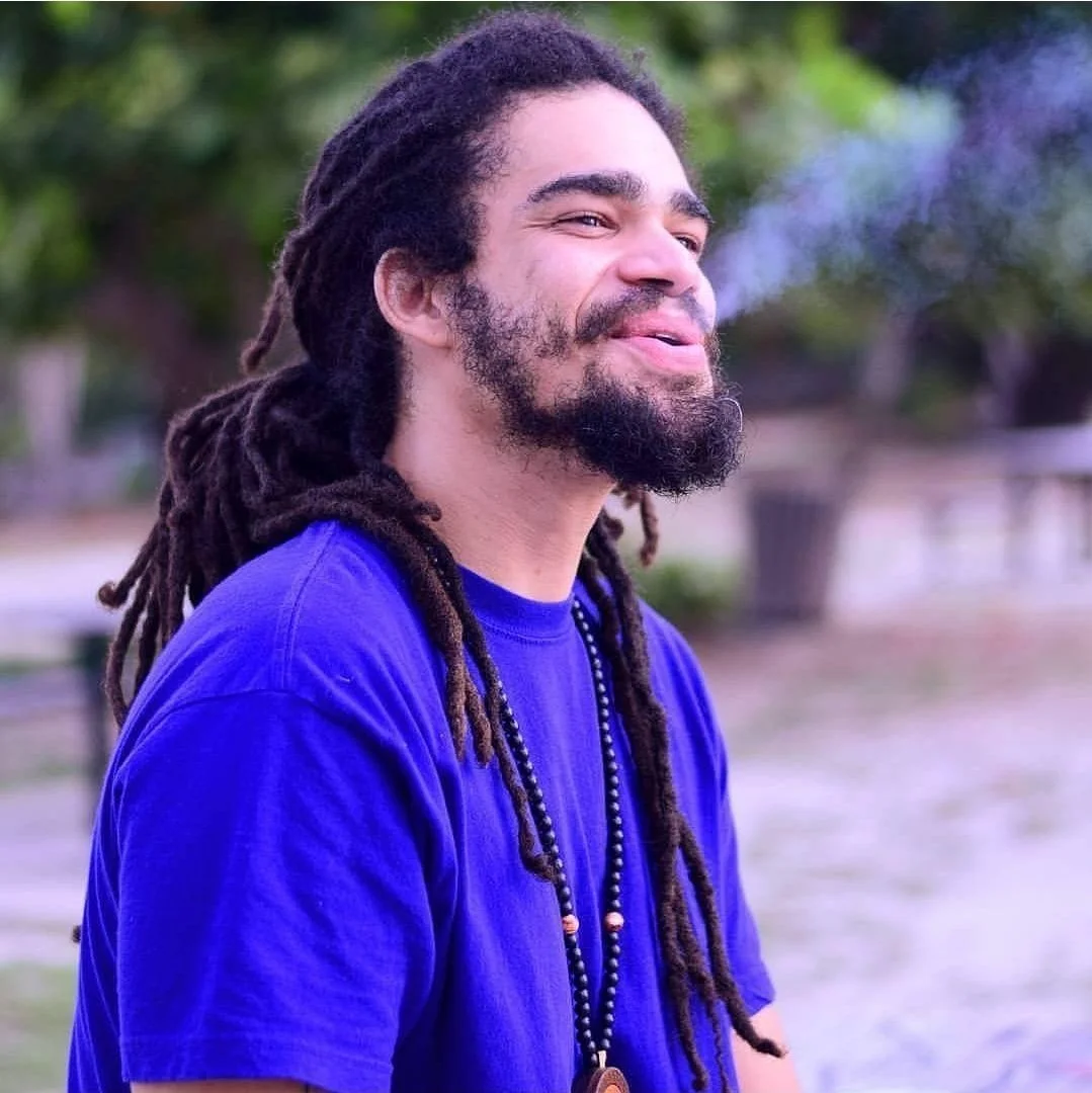 A man with dreadlocks and a beard wearing a blue shirt, smiling outdoors with trees in the background.
