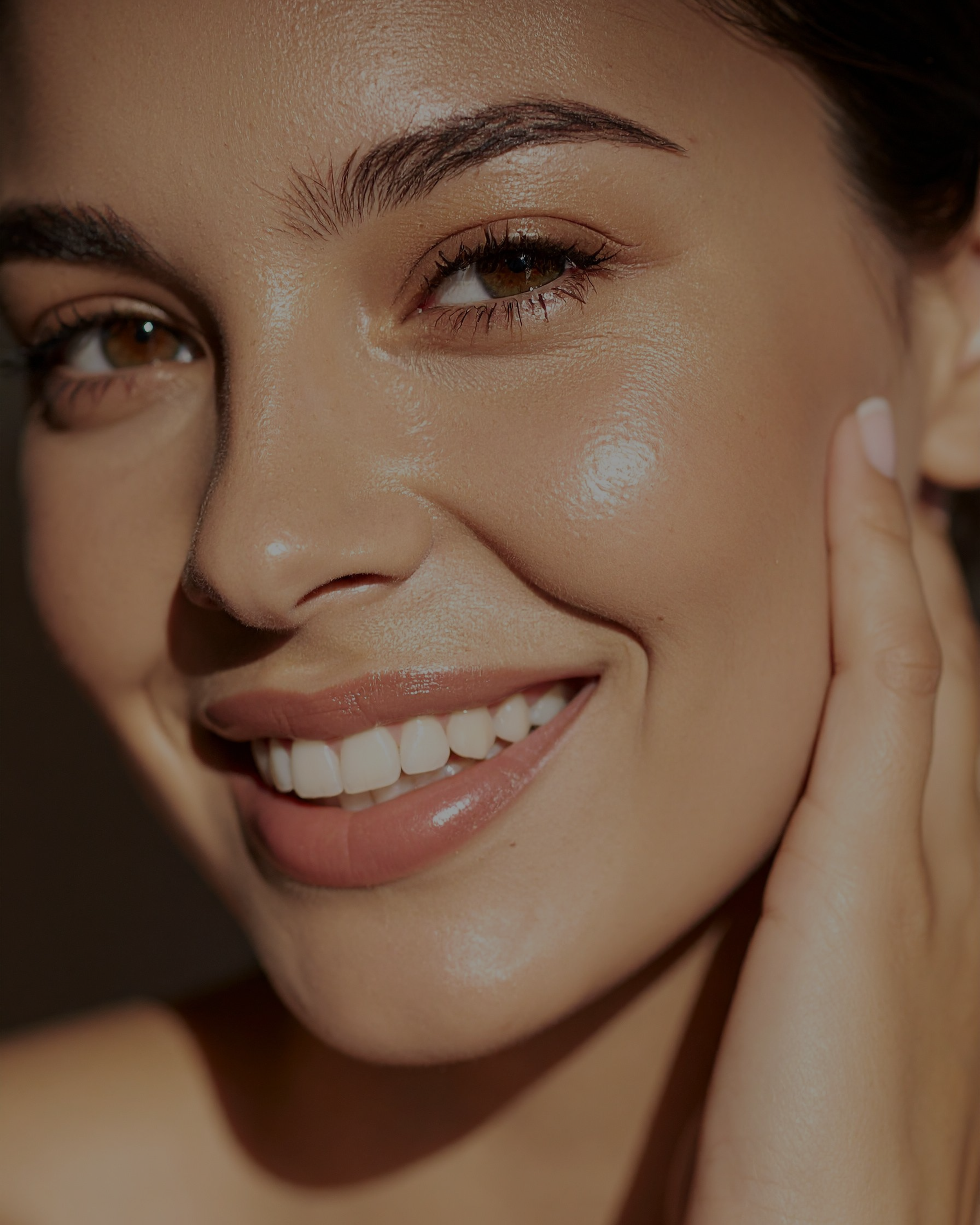 Close-up of a smiling woman with brown eyes, dark eyebrows, and smooth skin, touching her face gently with her hand.