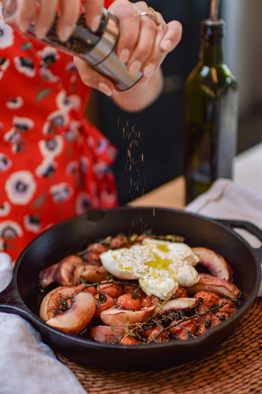Person seasoning a cast-iron skillet of roasted tomatoes, vegetables, and burrata with a pepper grinder