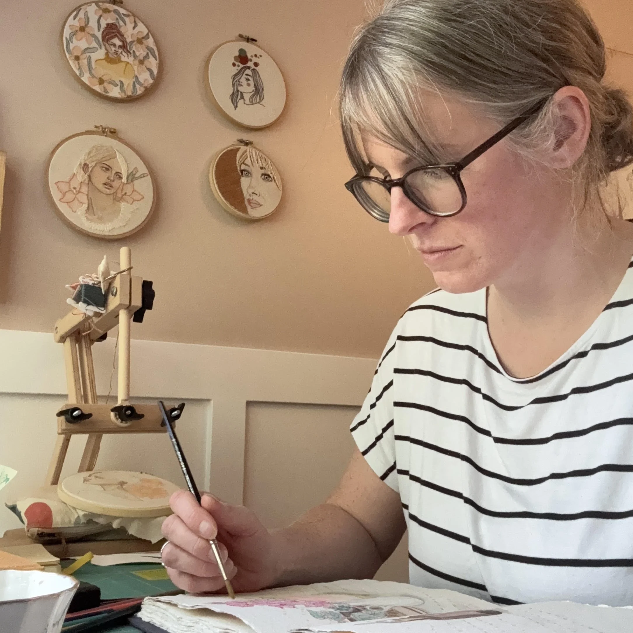 A woman with gray hair and glasses painting on fabric with watercolor paints. Behind her, on the wall, there are five embroidered portraits of women, each with different hairstyles and expressions.