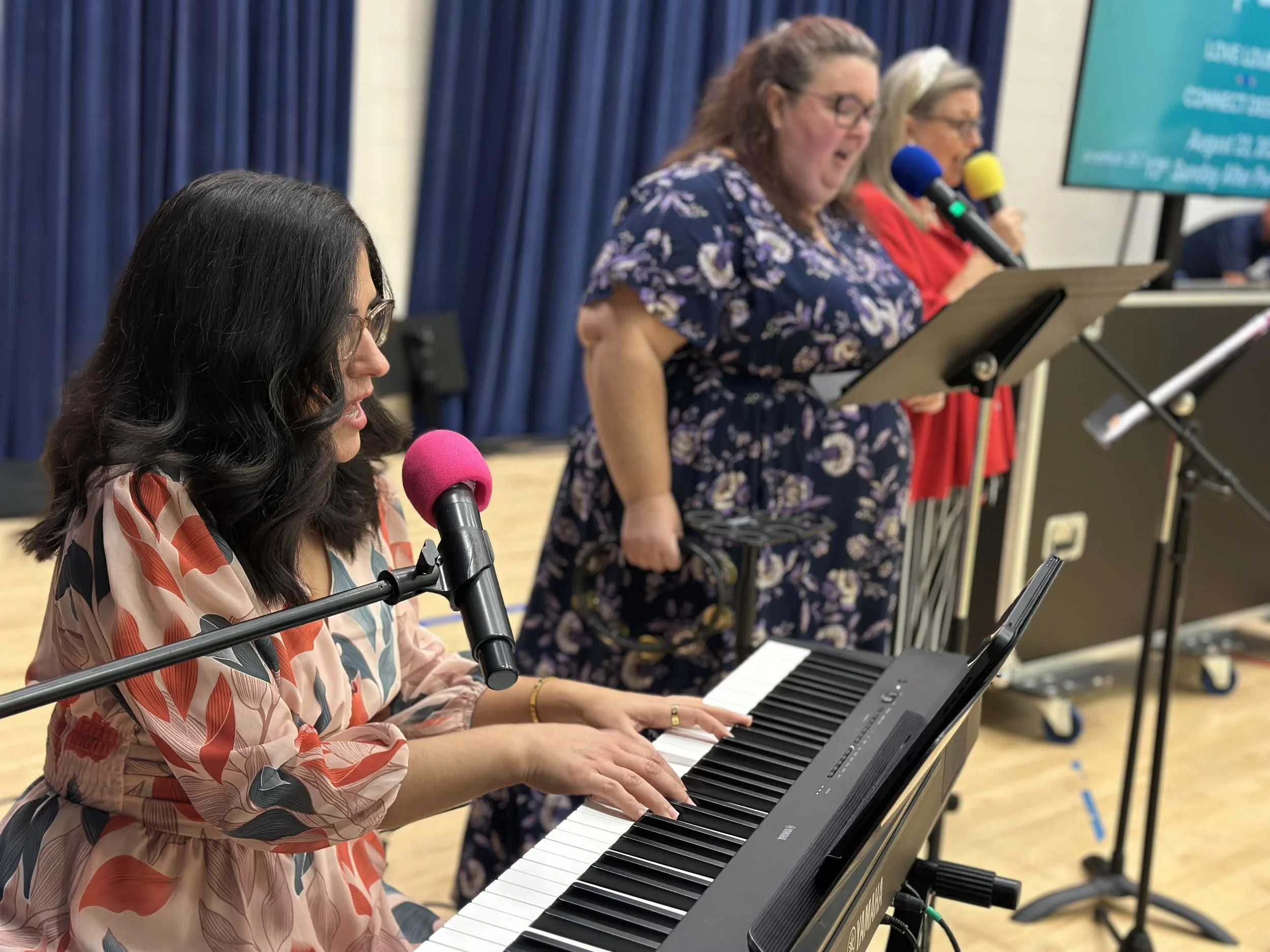 A woman with dark wavy hair and glasses playing a keyboard, with microphones placed in front of her. Behind her, other women are standing at music stands reading sheet music, speaking into microphones, in front of a dark blue curtain.