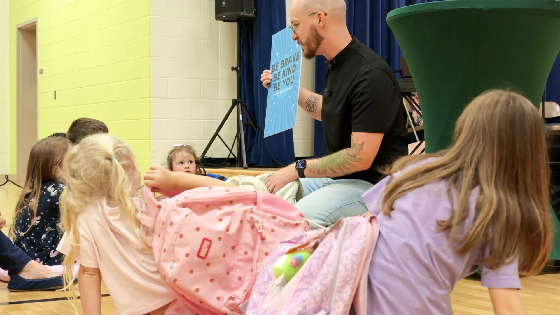 A bald man showing a poster to a group of kids. the poster says be brave, be kind, be you
