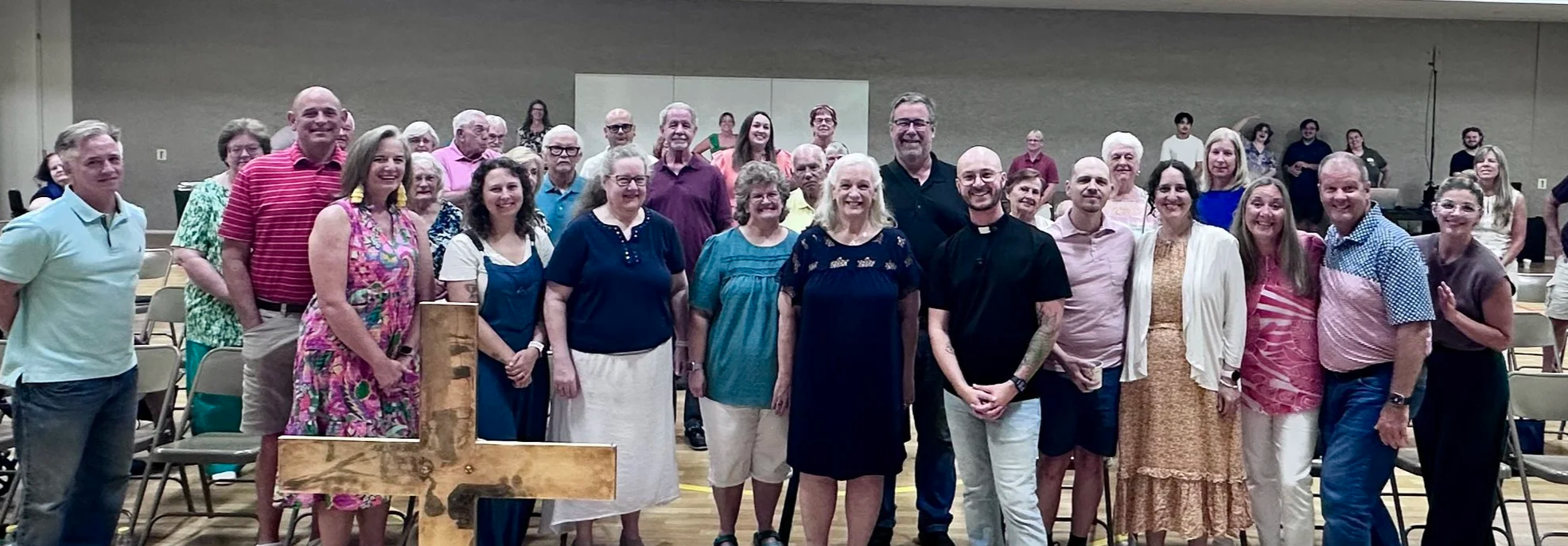 A group of people standing together in a church or community hall, smiling for a group photo. There are chairs behind them, and some people are seated in the background. The group includes men and women of various ages, some wearing casual clothing and others dressed more formally. A wooden cross is visible in the front left of the image.