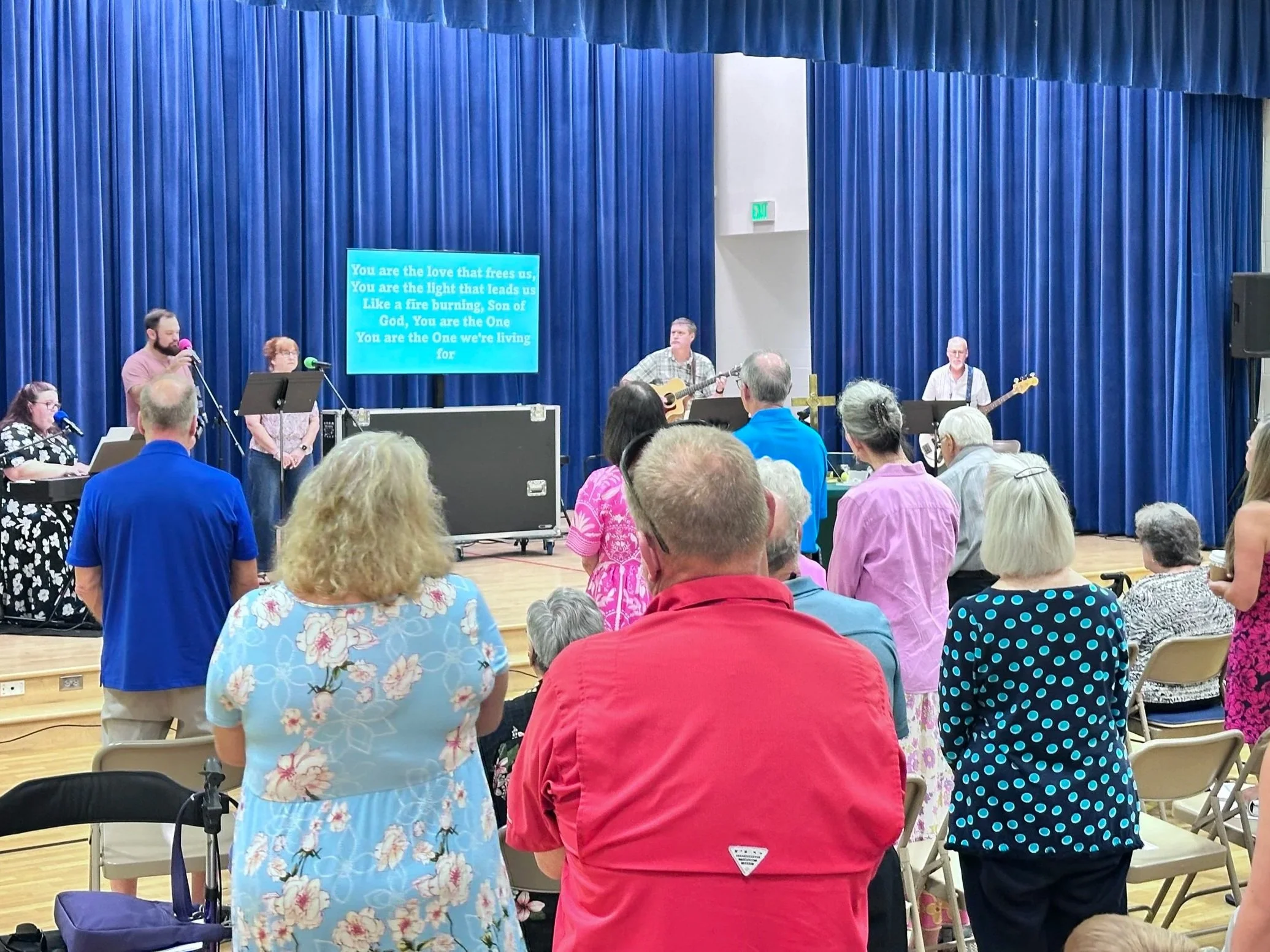 Group of people attending a religious gathering or choir performance in a hall with blue curtains. A few individuals are on stage with microphones and guitars. The audience is standing and facing the stage, some with head bowed or hands raised.