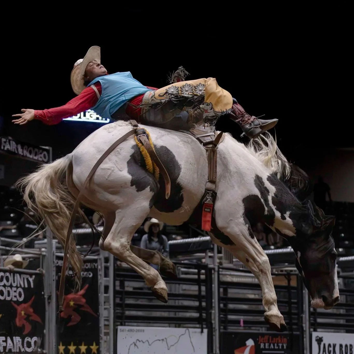 Person riding a bucking horse at a rodeo event, wearing a cowboy hat, rodeo attire, and the horse is mid-buck in an arena with banners and spectators in the background.