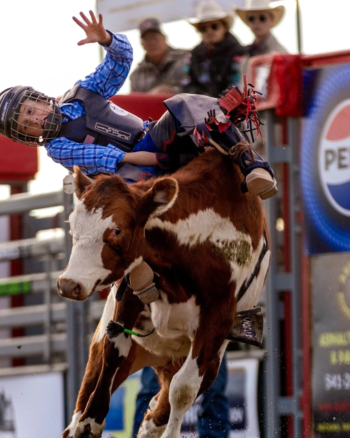 A young boy wearing a helmet, plaid shirt, and protective vest riding a cow during a rodeo event, with spectators in the background.