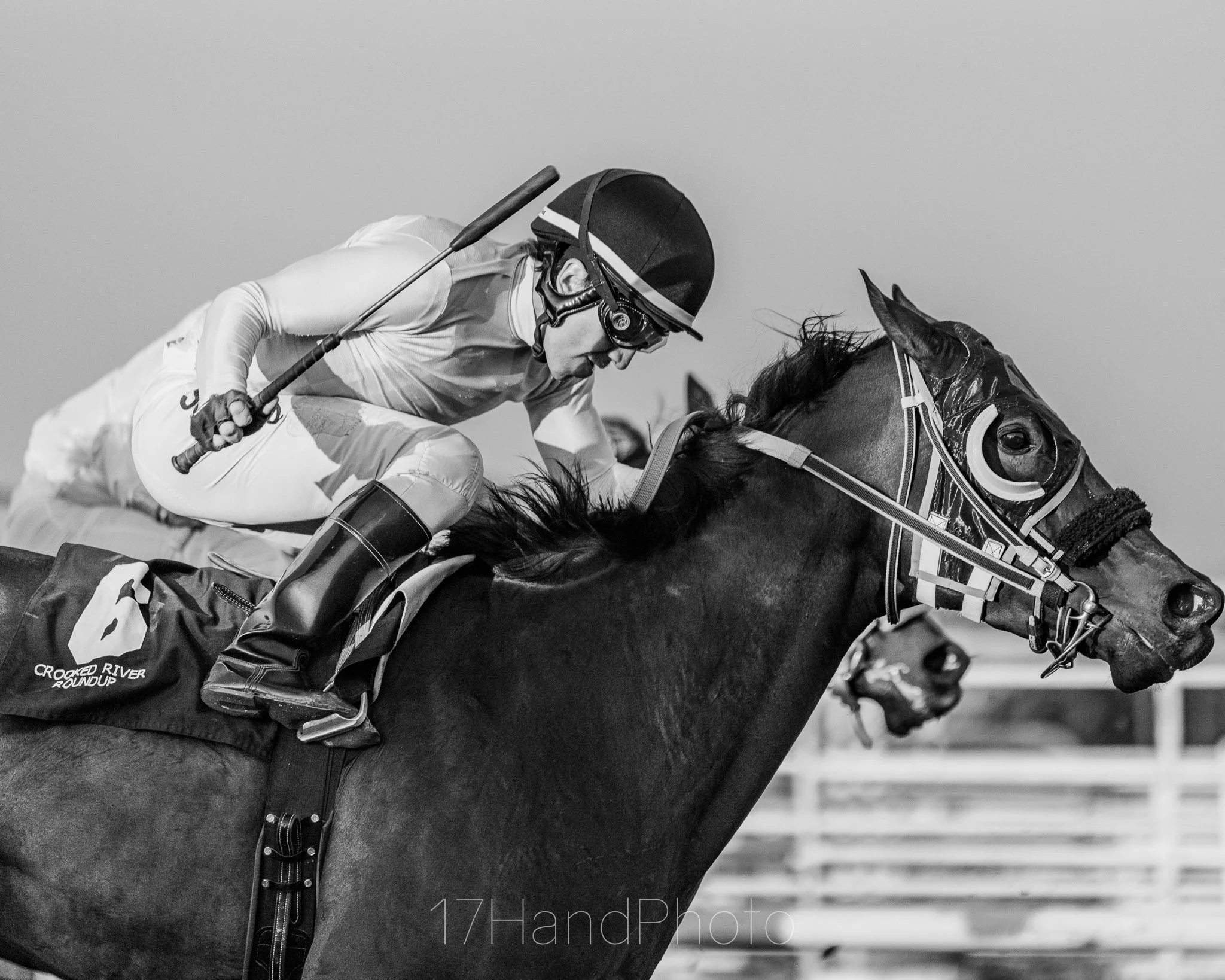 A jockey riding a horse during a race, with the jockey wearing protective gear including a helmet and goggles, holding a riding crop, in black and white.