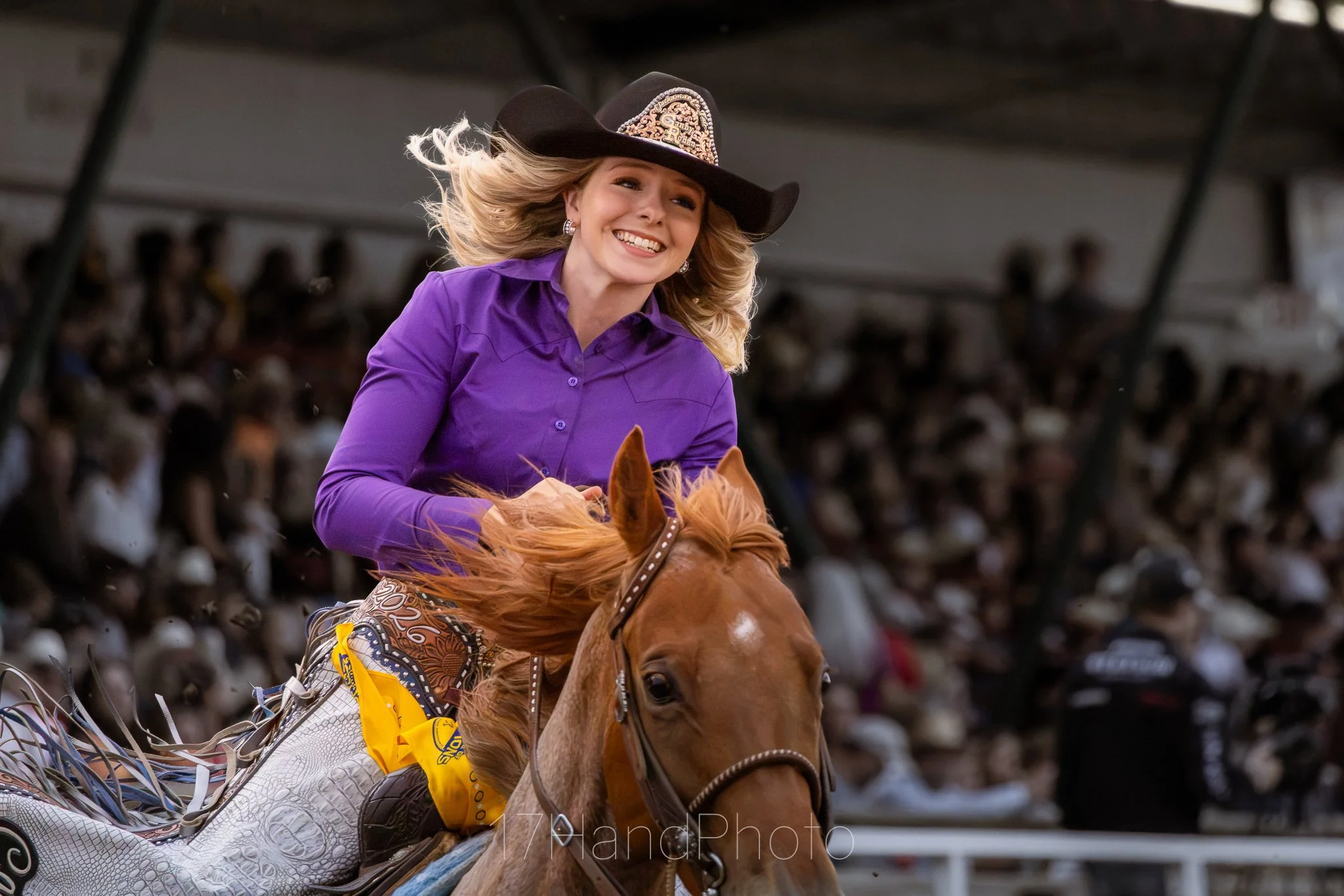 A woman wearing a black cowboy hat, purple long-sleeve shirt, and large earrings rides a brown horse. She has blonde hair flowing behind her and is smiling. The background shows an audience watching in a stadium or arena.